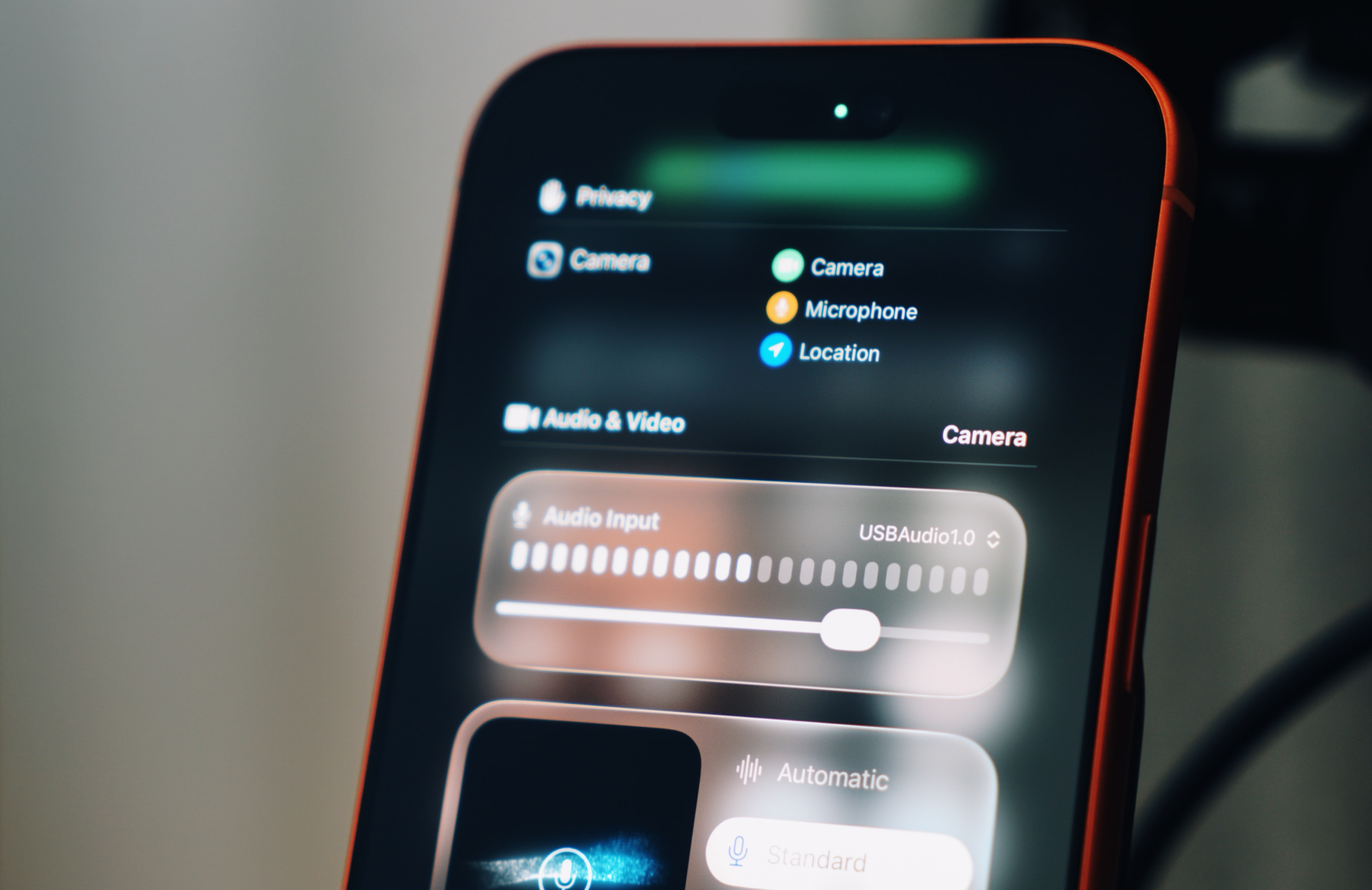Close-up of a smartphone screen displaying privacy settings, showing active Camera, Microphone, and Location usage icons, along with Audio Input settings with a volume slider. The phone has a black screen and a red border.

