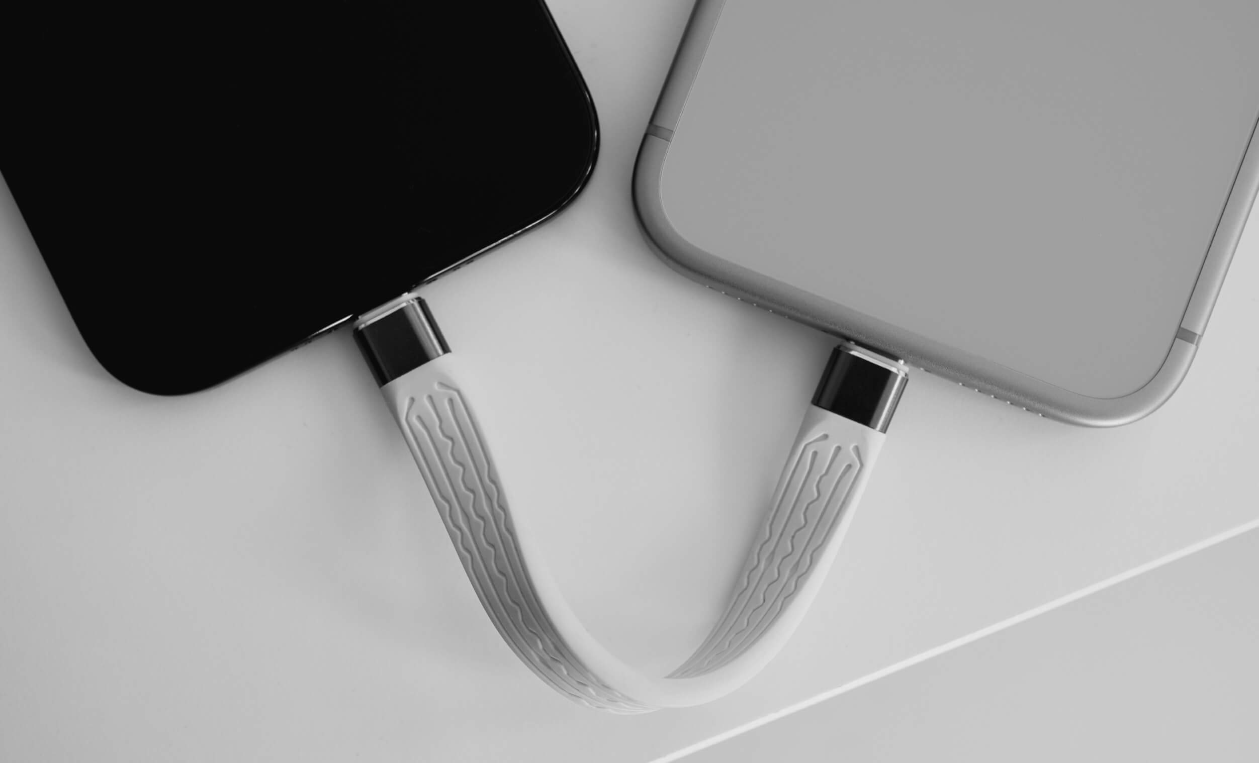 Two smartphones, one black and one silver, connected by a curved, patterned white charging cable on a white surface.