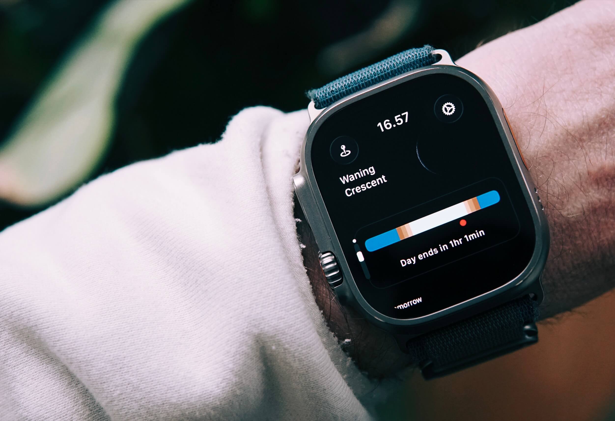 Close-up of a wrist wearing a smartwatch with a black screen displaying the time (16:57), location, and astrological data, including "Waning Crescent". Below is a graphic showing a day/night cycle, indicating the day ends in one hour and one minute. The watch is silver with a black strap, and the wrist is covered by a white sleeve.