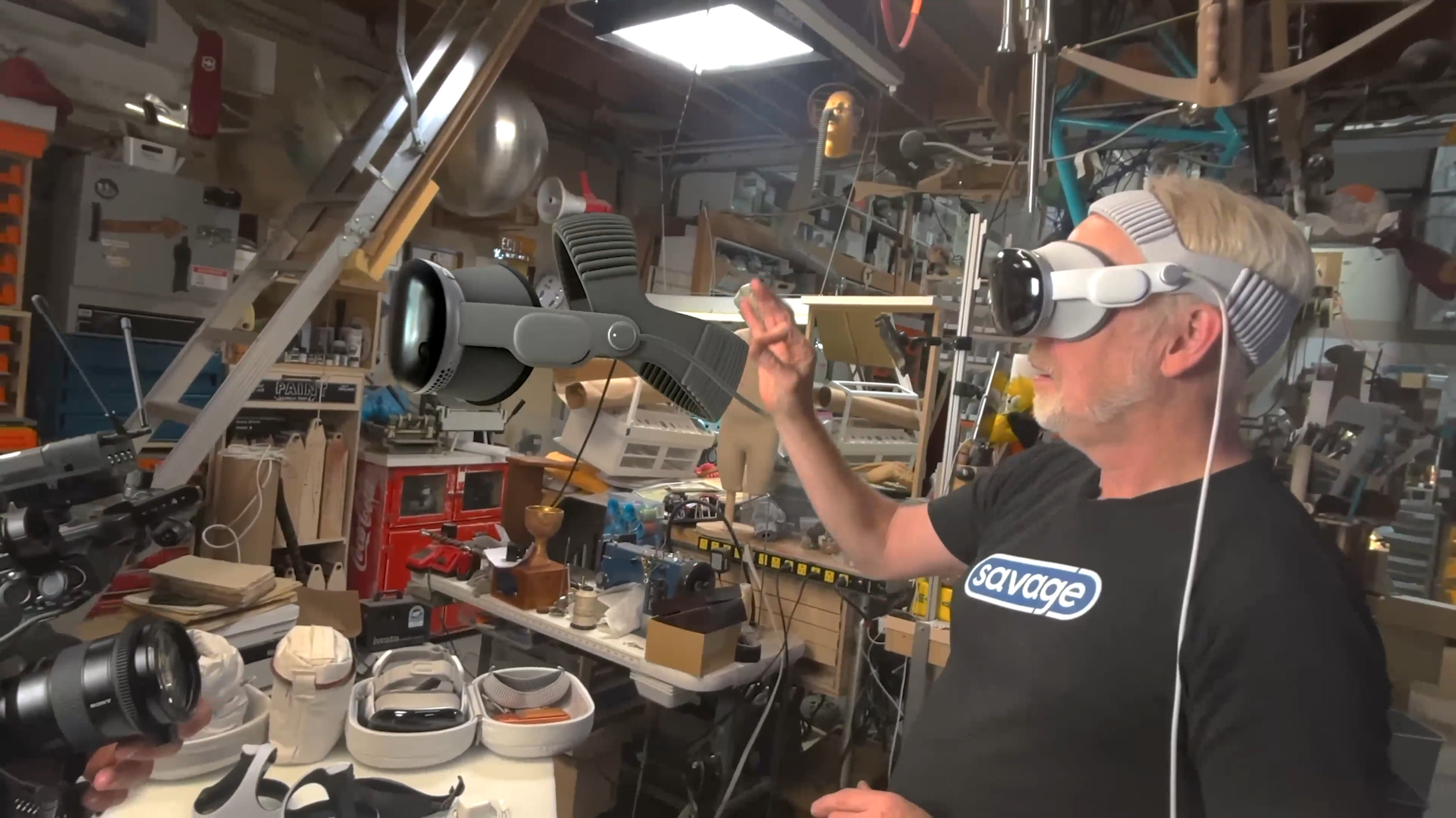 A man wearing a VR headset in a workshop, reaching out toward a 3D model of a headset floating in the air. The workshop is cluttered with tools and equipment, and the man's shirt has a "savage" logo.