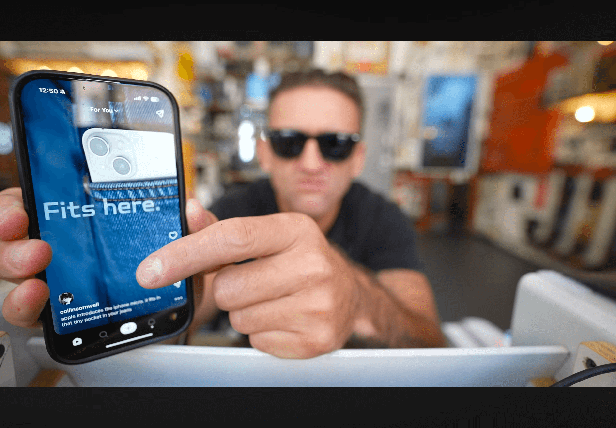 Man wearing sunglasses holds up a smartphone displaying a denim pocket with a white iPhone inside, with the text "Fits here" on the screen, demonstrating the phone's size.
