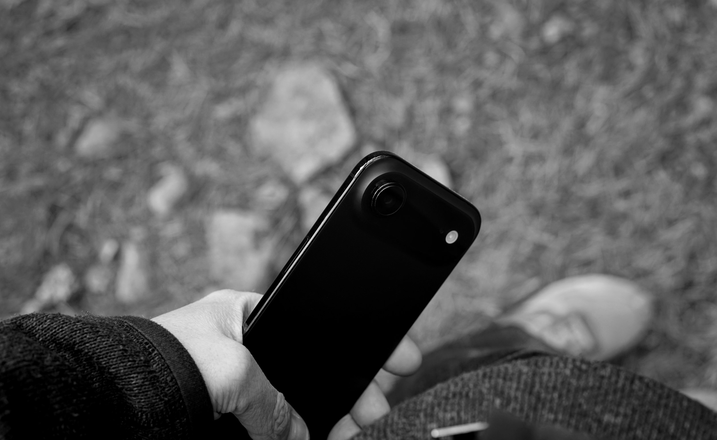 Monochrome photo showing a person holding a black smartphone, with the phone's camera lens visible, the background is blurred showing grass and stone.