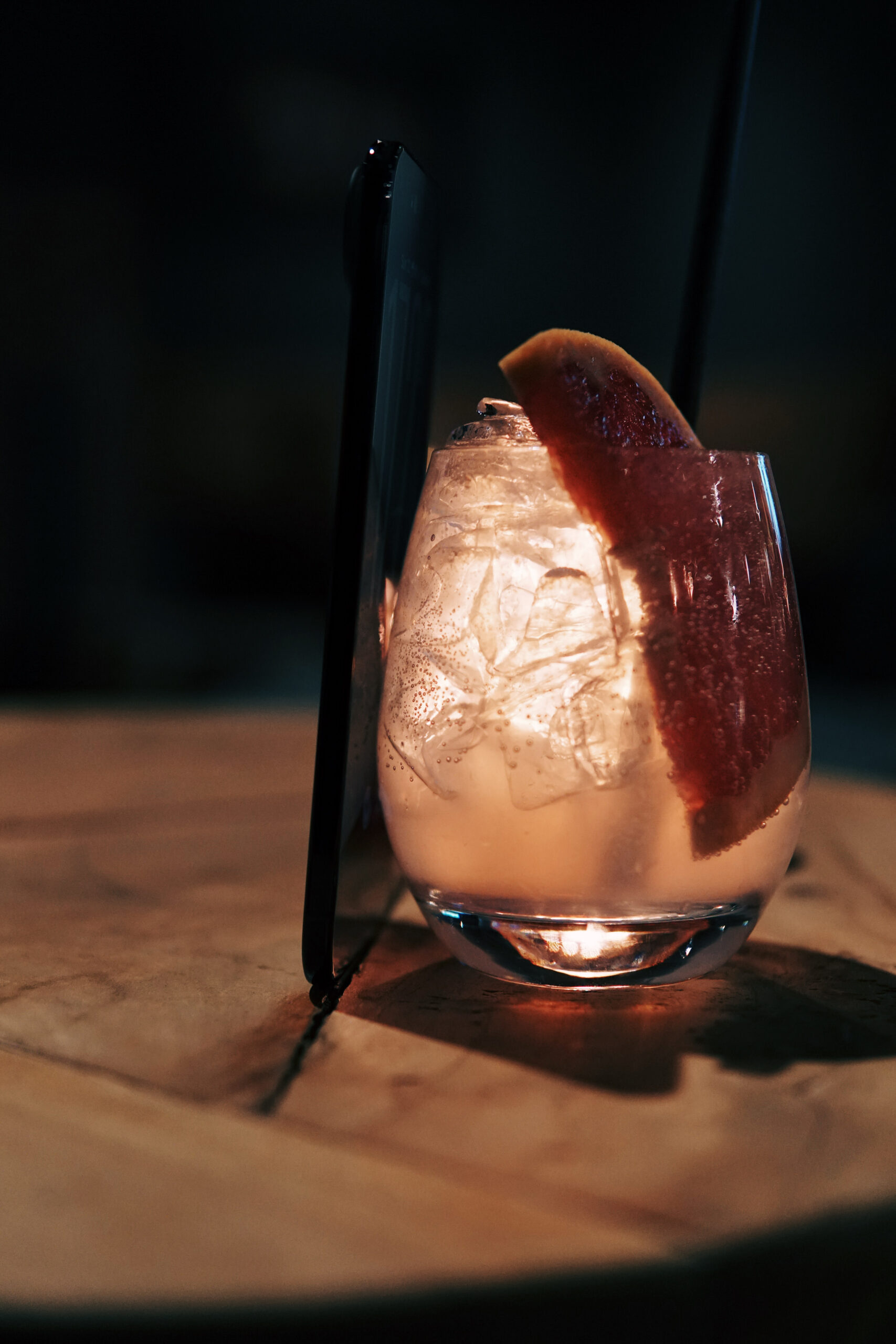 Stemless cocktail glass with a pale pink drink containing ice cubes, a grapefruit slice, and a black straw, resting on a wooden surface with a dark, blurred background.