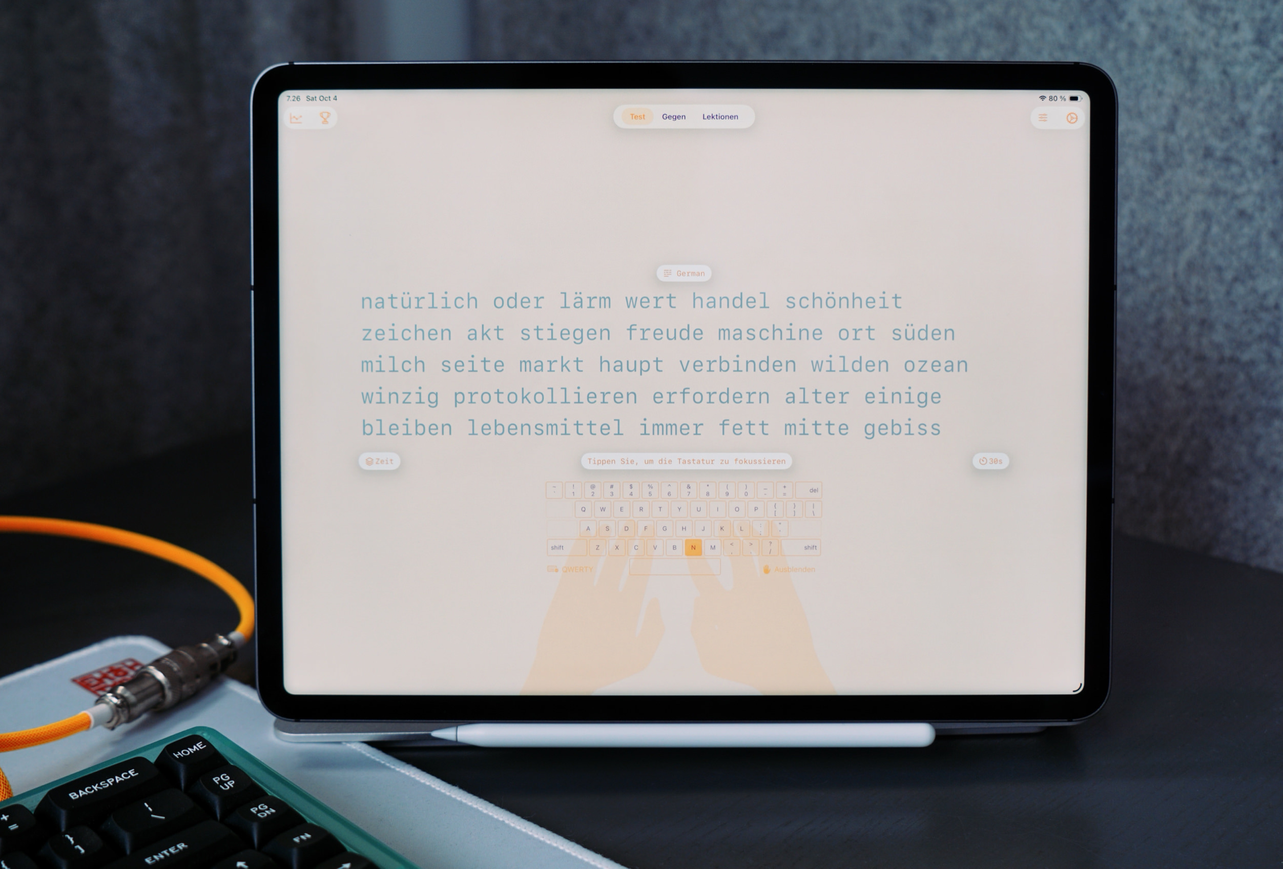 An iPad displays a text-based German lesson on its screen, with a keyboard visible at the bottom, a stylus below the screen, and a mechanical keyboard and cabling in the foreground.