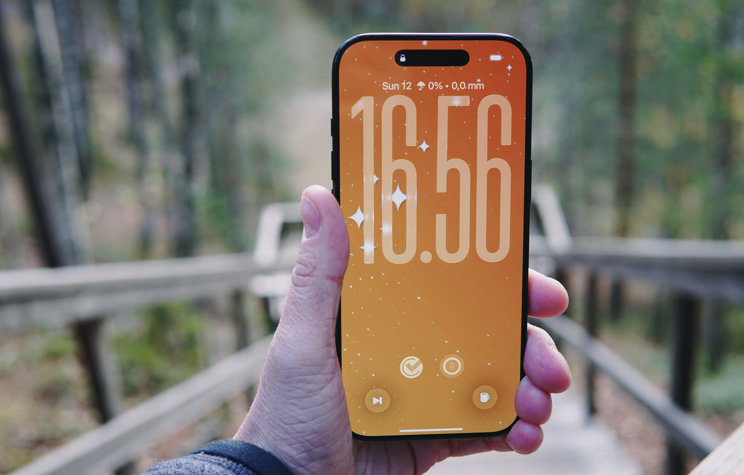 A hand holding a smartphone displaying a bright orange digital clock displaying 16:56. The background is of a blurred out wooden structure with trees.