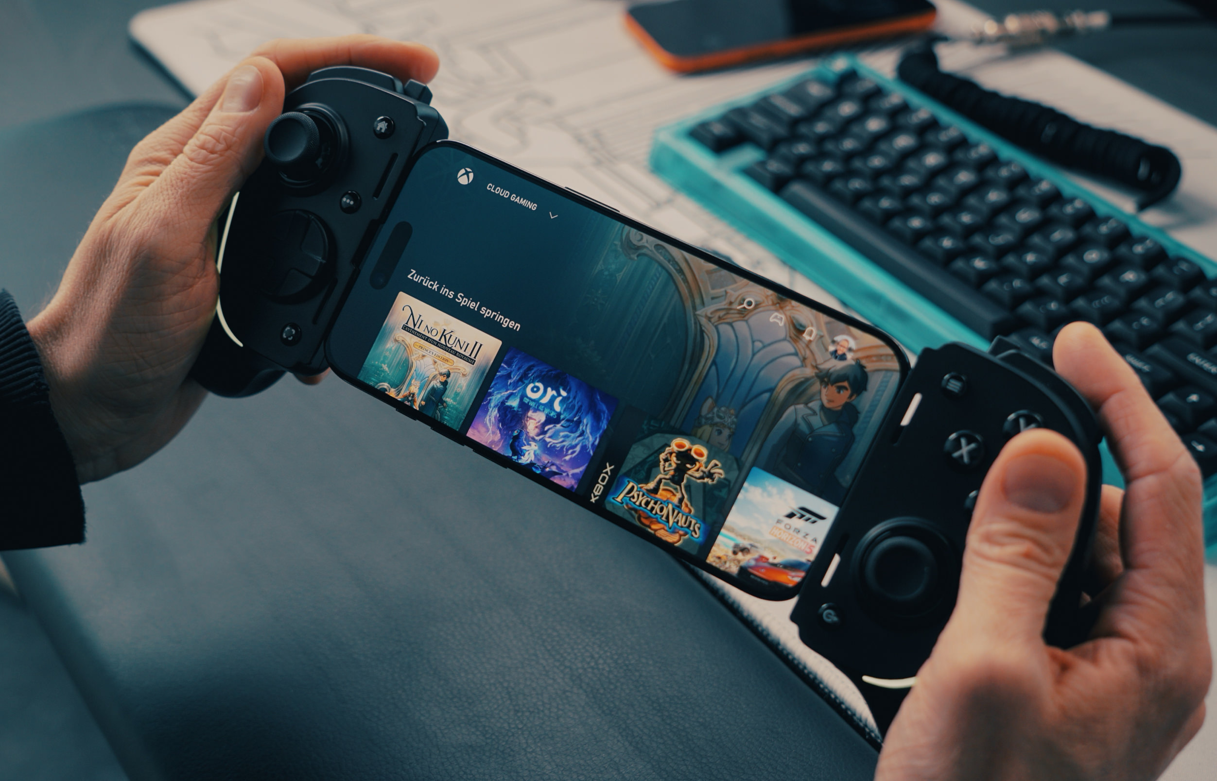 Person holding a smartphone in a black controller, displaying the Xbox Cloud Gaming interface with game thumbnails like Ni No Kuni II, Ori, Psychonauts, and Forza Horizon. A turquoise keyboard and a smartphone are in the background.
