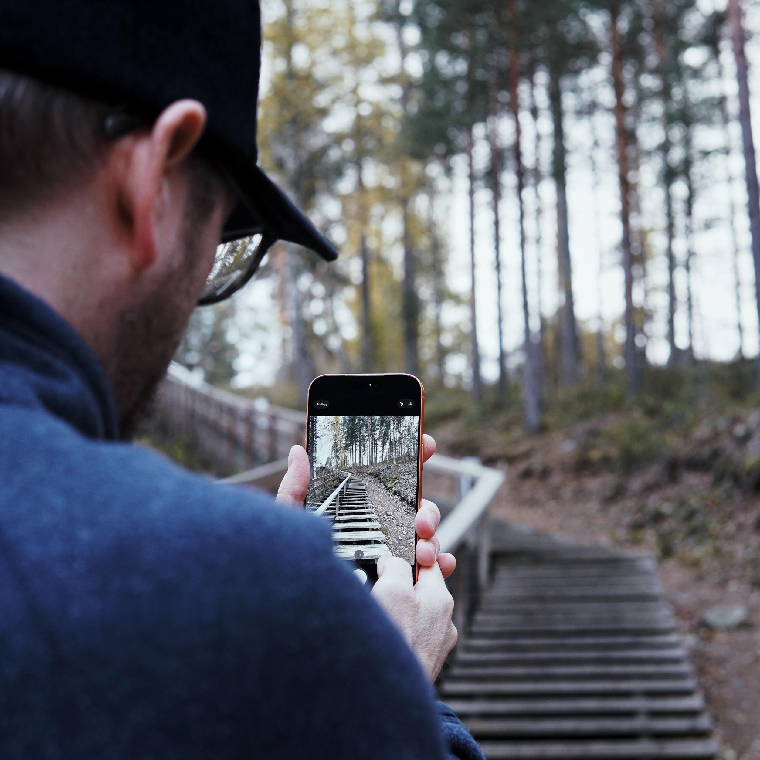 A person, wearing a black cap and glasses, holds a smartphone capturing a photo of a wooden staircase winding through a forest with tall trees. The smartphone screen displays the staircase and surrounding forest, and its own photo of the scene.