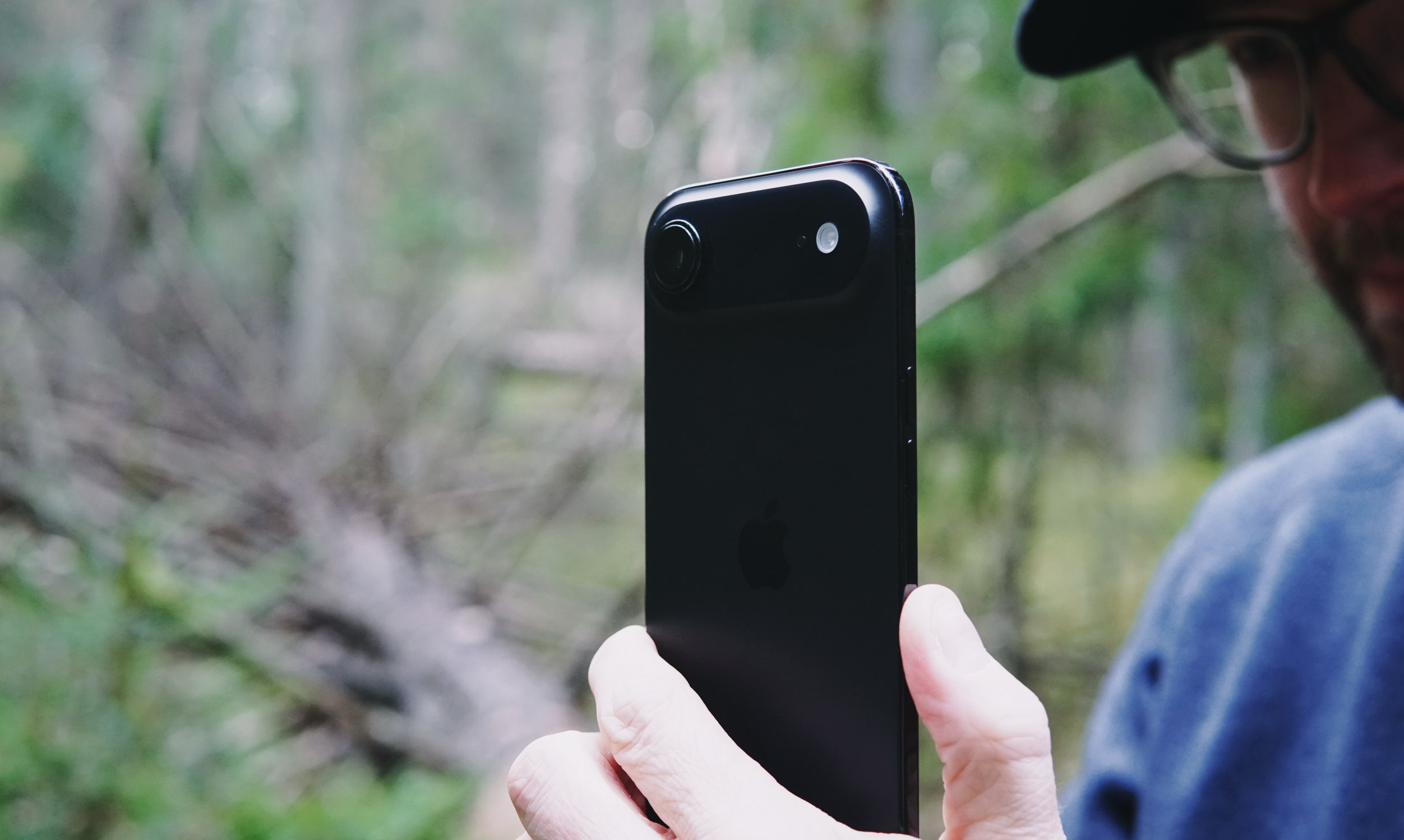 Close-up of a person holding a black iPhone in front of a blurry forest background. The phone's camera and flash are visible, along with the Apple logo. The person is wearing glasses and a blue shirt.