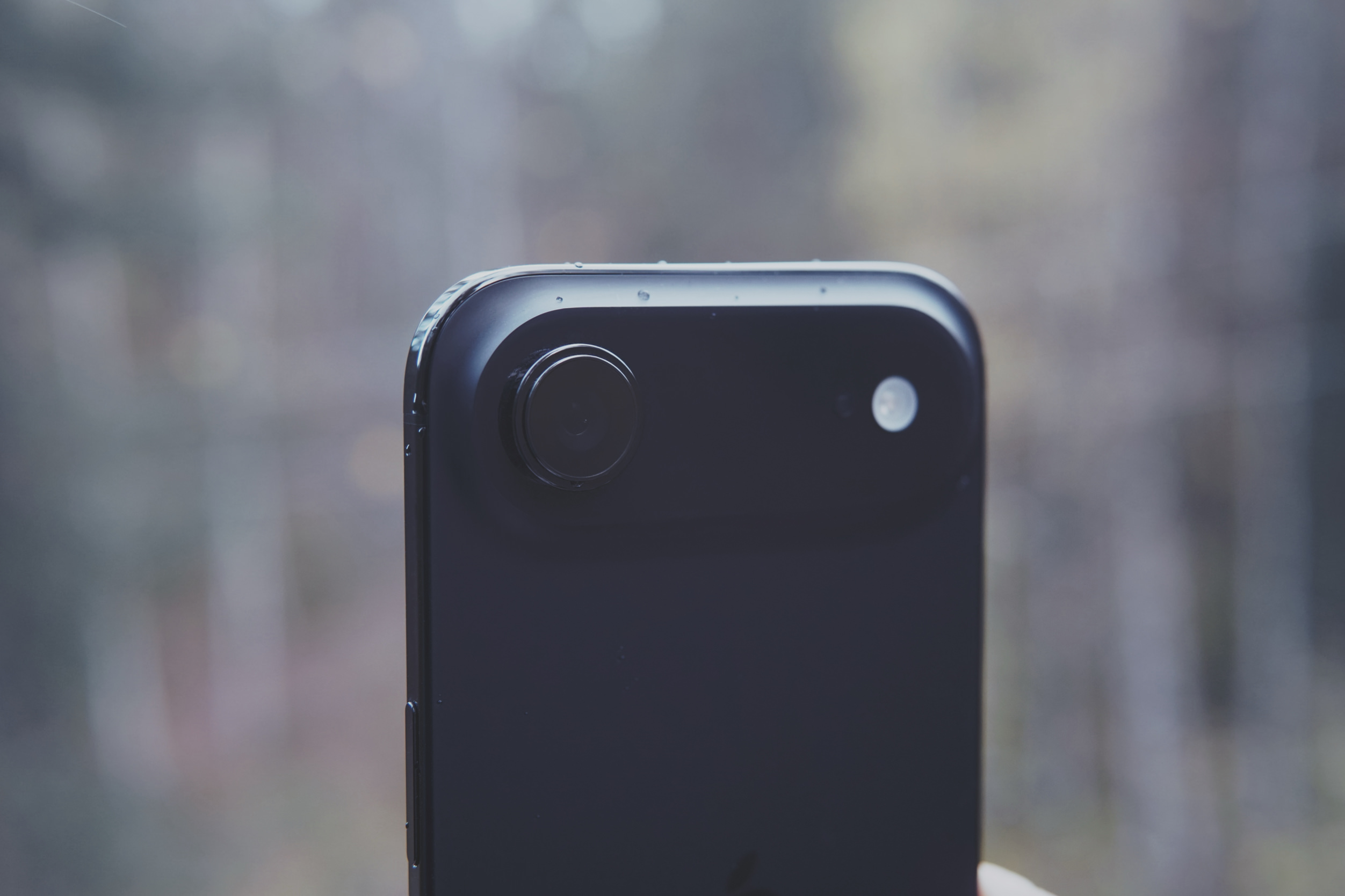 Close-up shot of the top portion of a black smartphone with a single, central, circular camera lens and an LED flash. The phone's surface has small water droplets. The background is blurred, suggesting an outdoor setting.