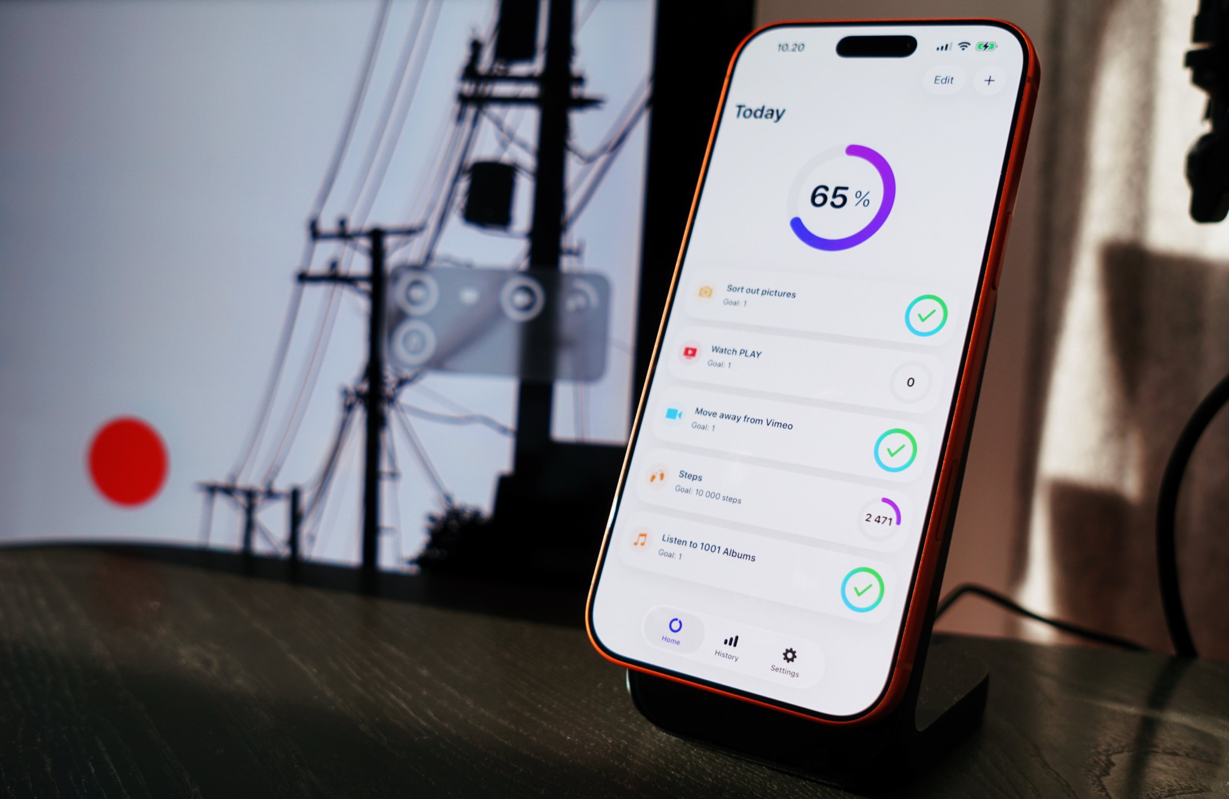 A smartphone displaying a productivity app, positioned on a dark desk with blurred out background elements; the phone is on a black stand showing today's progress as 65%, with tasks listed below.