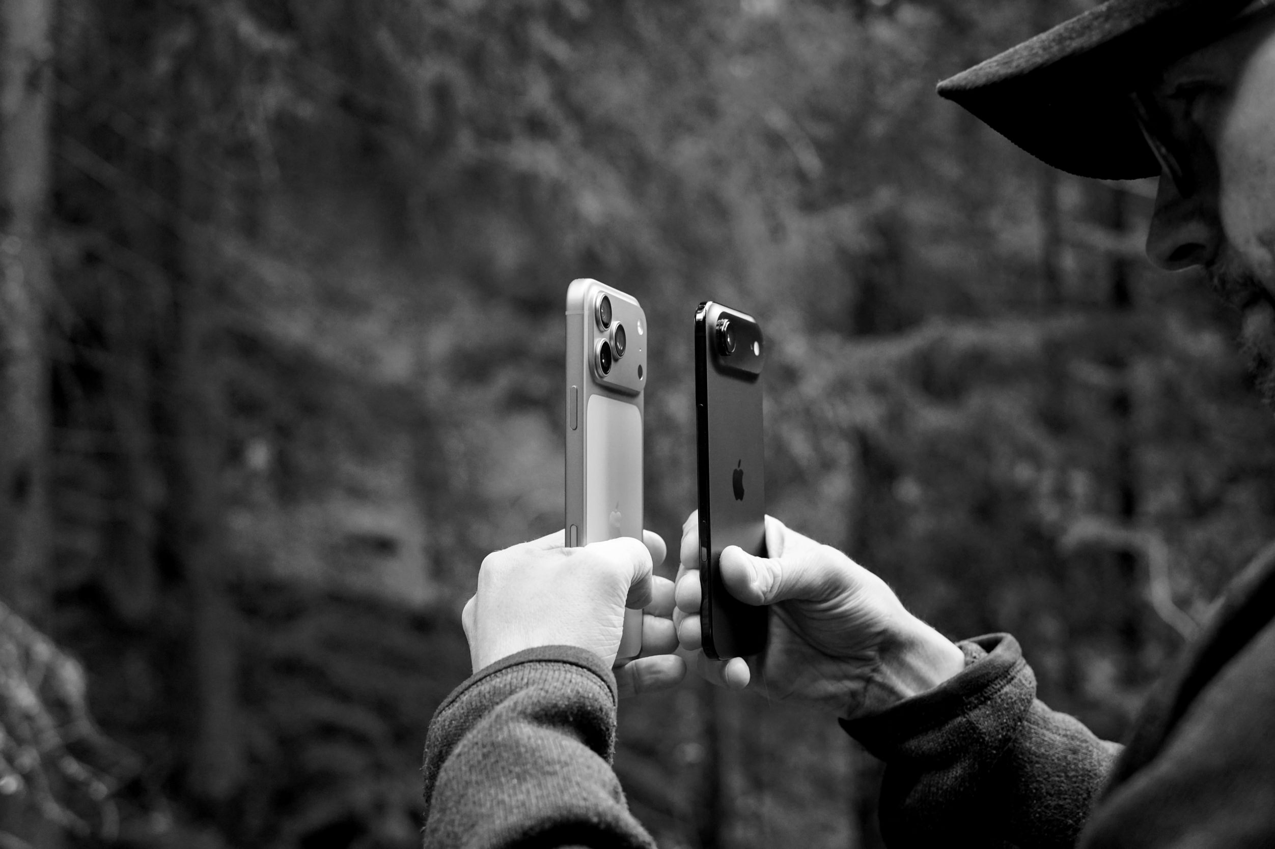 A black and white photo of a man holding two Apple iPhones, one silver and one black, displaying their cameras against a blurry forest background. The man, only partially visible, wears a dark jacket and hat. The silver iPhone shows three lenses, while the black one has a single lens.