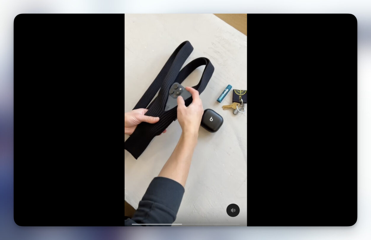 A close-up shot of a person's hands placing a smartphone into a black, ribbed, wearable pouch. The pouch, designed to be worn around the neck, has a long strap folded over and is positioned on a light gray surface. Alongside the pouch are various items, including a black earbud case with the number "6" printed on it, a "BigFix" branded lip balm, a key ring with keys and a “Stone Island” tag, and a small key. The person's arm, visible at the bottom of the frame, is wearing a black long-sleeve shirt. The scene is framed as a video in a wide aspect ratio, with black borders on the top and bottom.