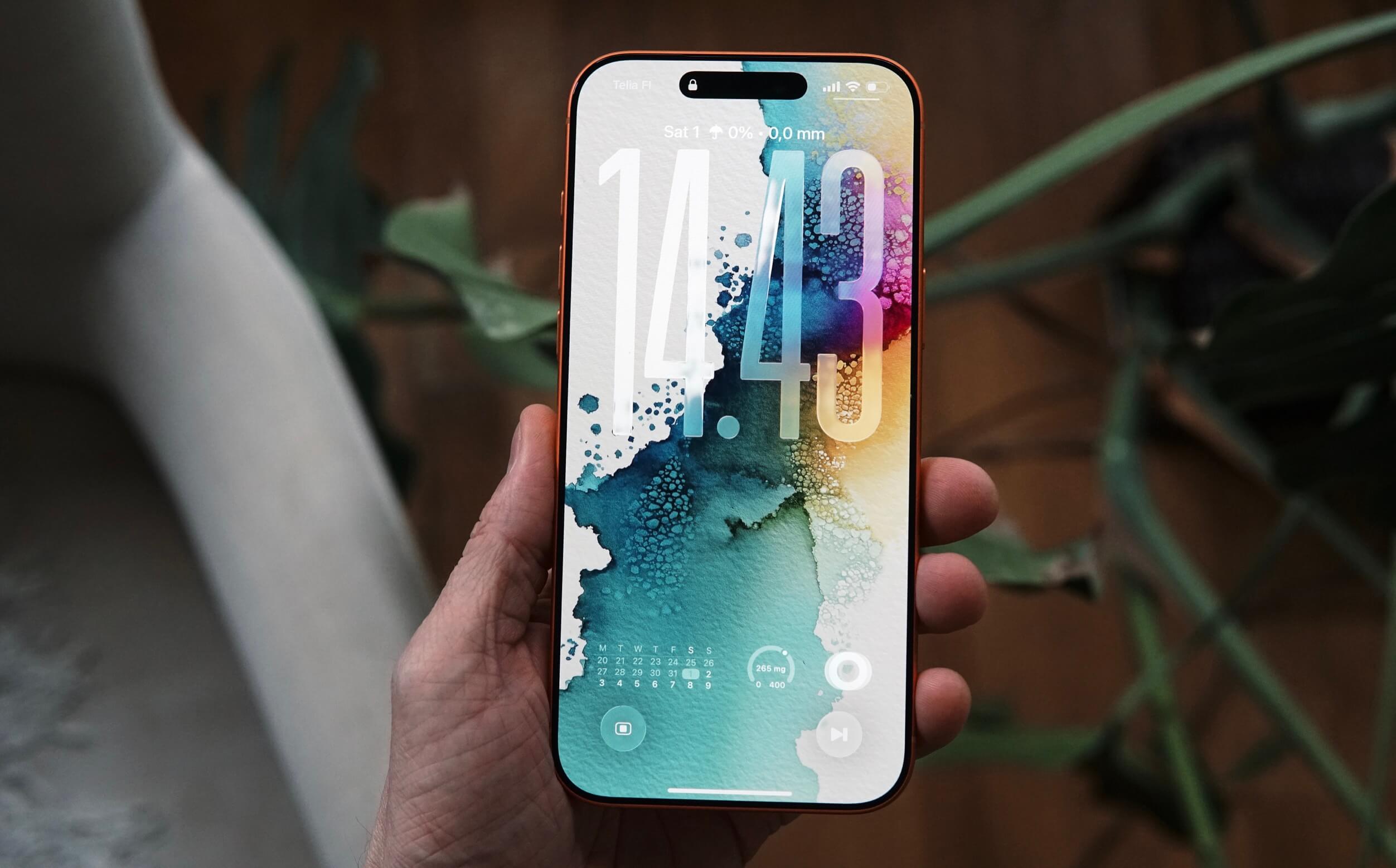 A hand holding a modern smartphone with a colorful abstract watercolor wallpaper displaying the time "14:43" with calendar and fitness widget on a blurred background of plants and a wooden surface.
