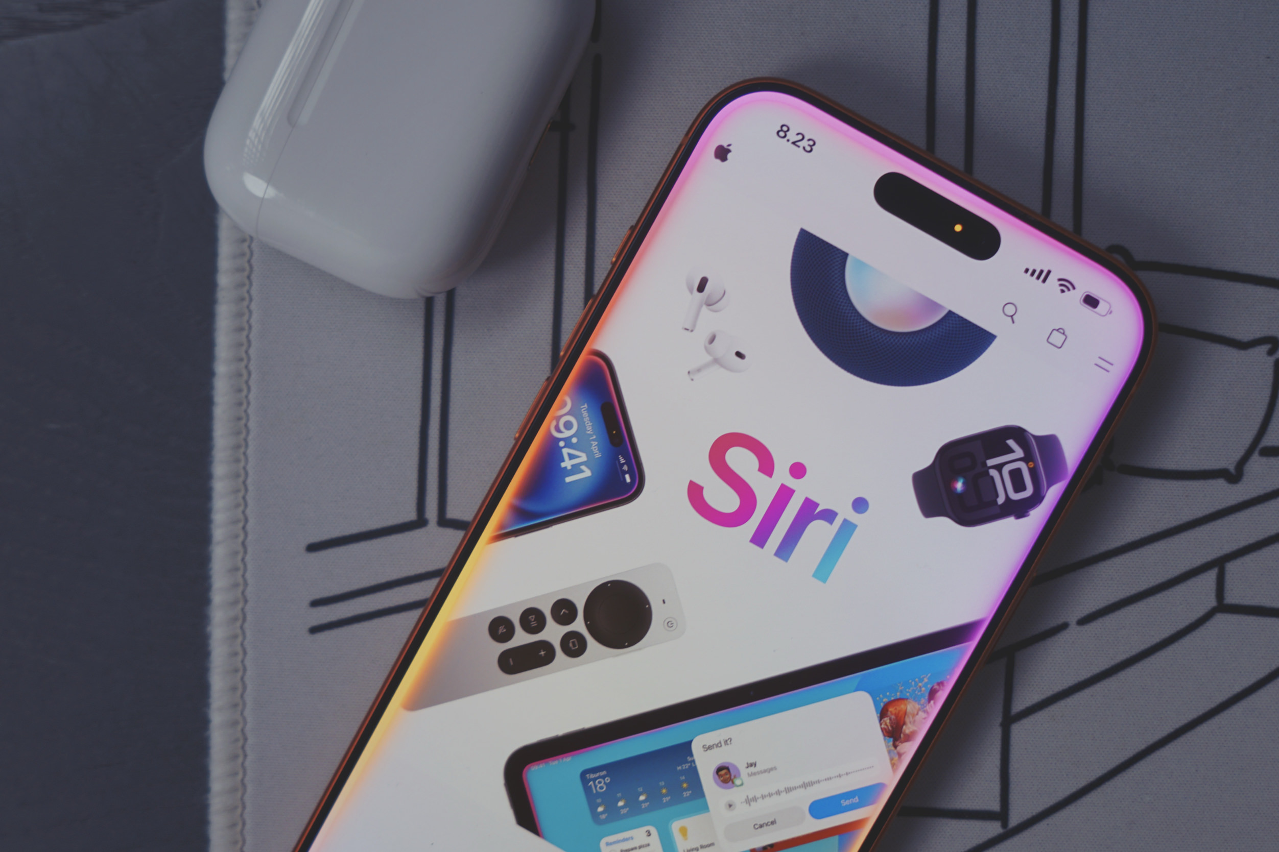 Close-up shot of an Apple iPhone displaying a Siri interface, various Apple products, and the time “8.23” in the top left corner of the screen. The screen is light-colored and features a large “Siri” logo. Surrounding the “Siri” logo, a pair of white Apple AirPods, a smartwatch with the number “10” on the screen, a remote control with a circular pad in the center, and a display with the temperature of “18°.” Additional screens on the iPhone display “Tuesday 1 April 09:41.” In the bottom of the screen, there is a display including “Send it?” and the name “Jay” with “Send” and “Cancel” buttons. A white Apple AirPod case rests beside the phone. The phone and AirPods case rest on a grey patterned surface.