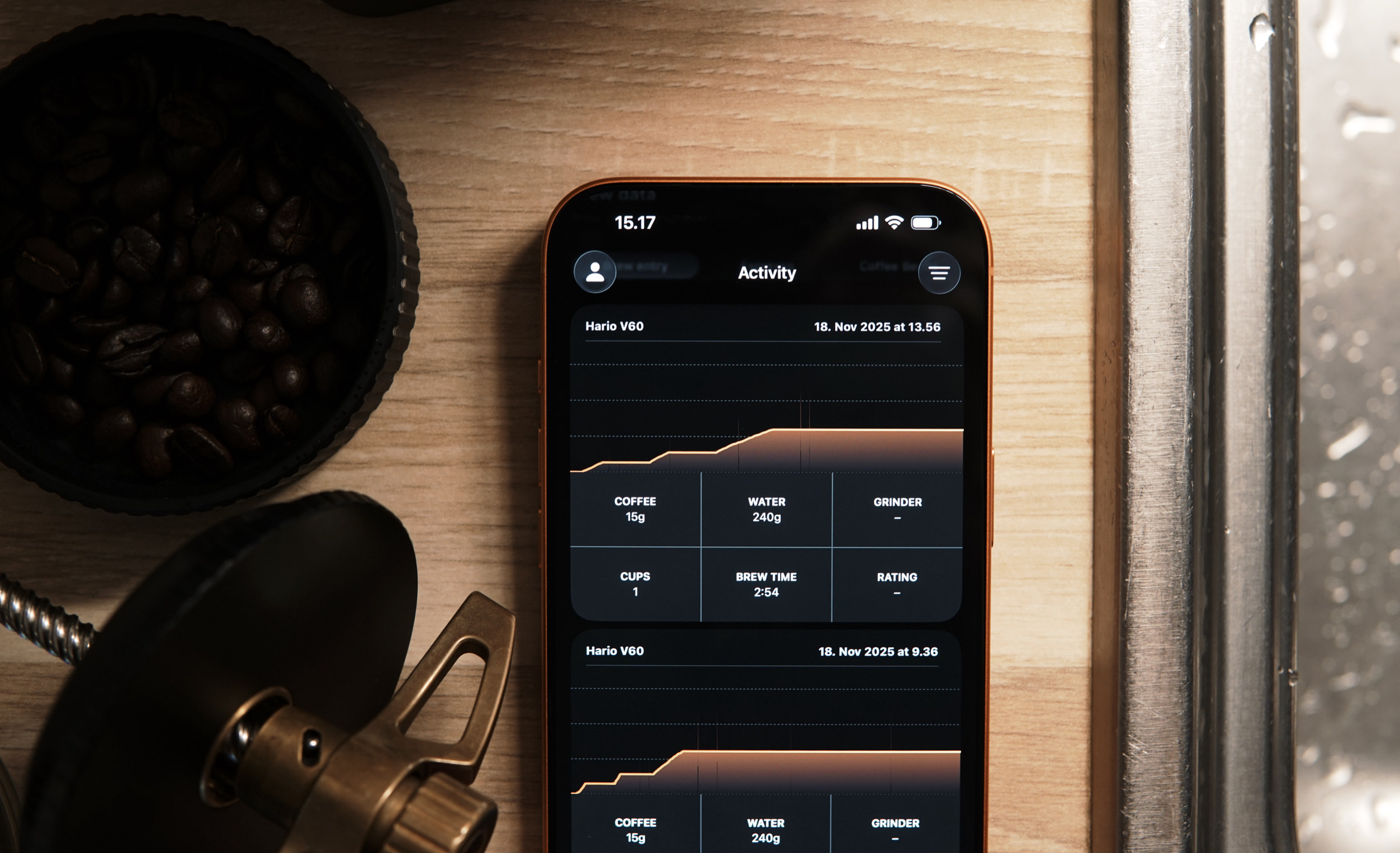 A smartphone displaying a coffee brewing activity log is laid on a light wood surface. The top portion of the phone displays "15.17" and the word "Activity". Below the title, the activity log displays, "Hario V60" and the date "18. Nov 2025 at 13.56". The log displays a graph with additional data points below it: "COFFEE 15g", "WATER 240g", "GRINDER", "CUPS 1", "BREW TIME 2:54", "RATING -". Beneath this section, is a second log showing the same details, but a different date. A coffee grinder and a bowl of coffee beans lay to the side. The surface is next to a metallic kitchen fixture.