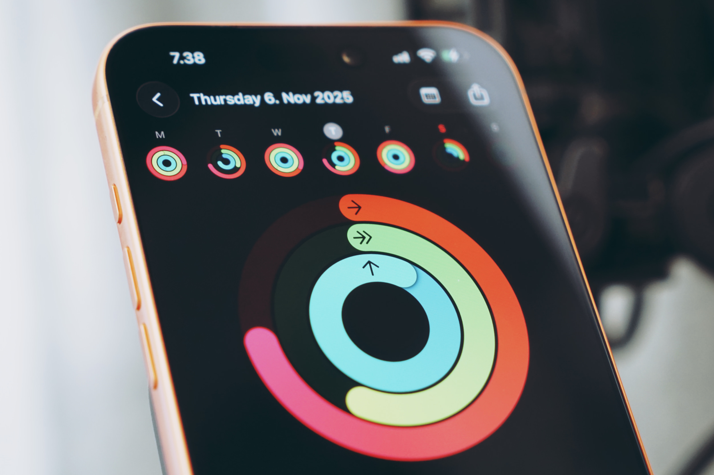 A close-up shot of a smartphone screen displaying an activity tracker app. The time at the top of the screen is "7.38" and the date "Thursday 6. Nov 2025" is shown below that. The app displays fitness rings, with three colored rings: pink, green, and blue, representing activity goals. The pink ring is partially filled, the green ring is mostly filled, and the blue ring is at least halfway filled. There is a "back" arrow at the top left of the screen and various notification icons at the top right.  The phone has a gold-colored frame, and the background is blurred.