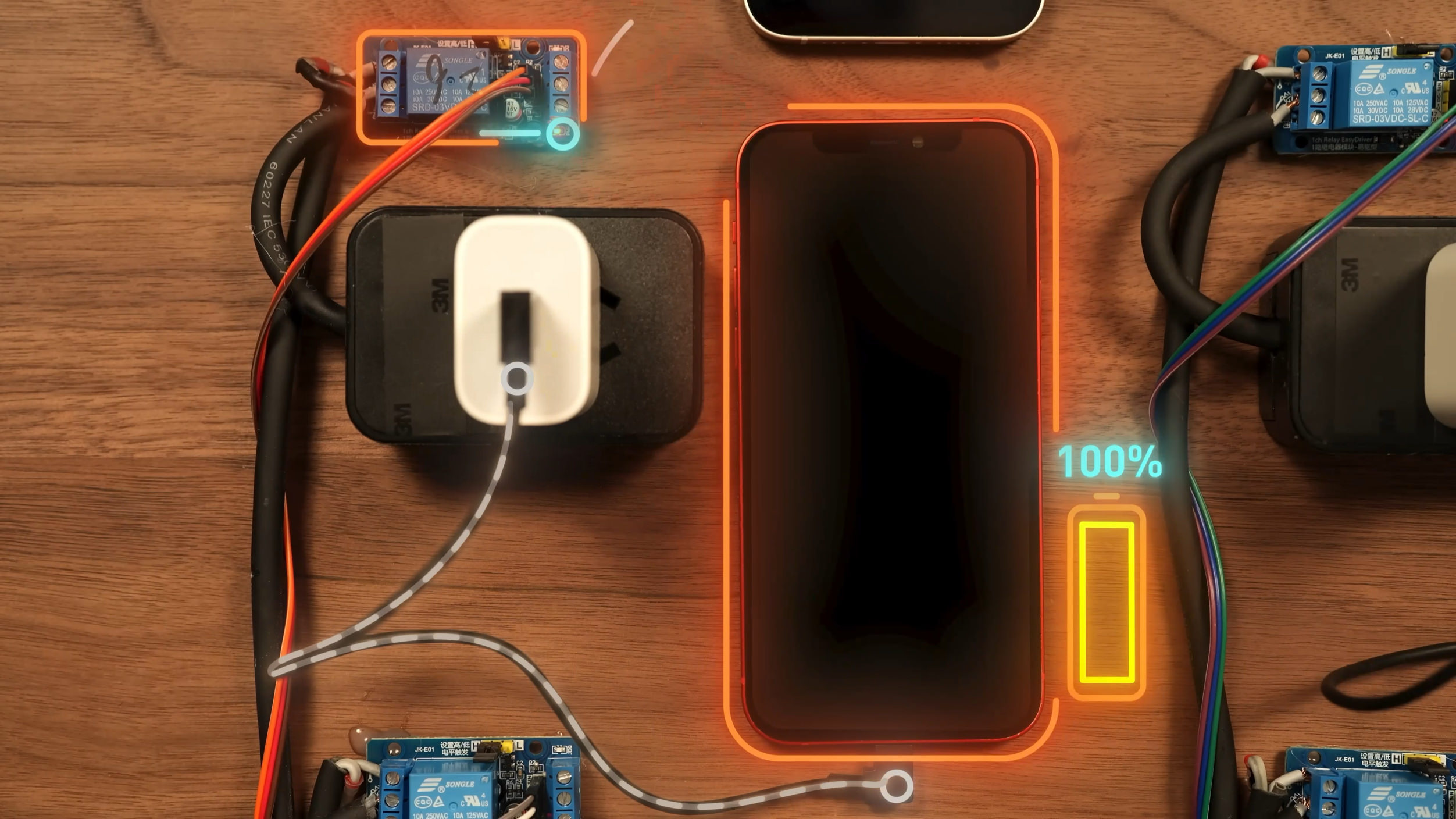 Overhead shot of a wooden table with electronic components and a smartphone. The main subject is a black smartphone with an orange glowing outline, with a graphic displaying "100%" and a battery charging icon. The phone is connected to a white power adapter plugged into a black box, with wires running to circuit boards with the label “SONGLE”, and the model is SRD-03VDC-SL-C. Other text on the components includes “10A 250VAC 10A 125VAC”, “JK-E01”, “1ch Relay EasyDriver”, and "NVINY 60227 IEC 530".
