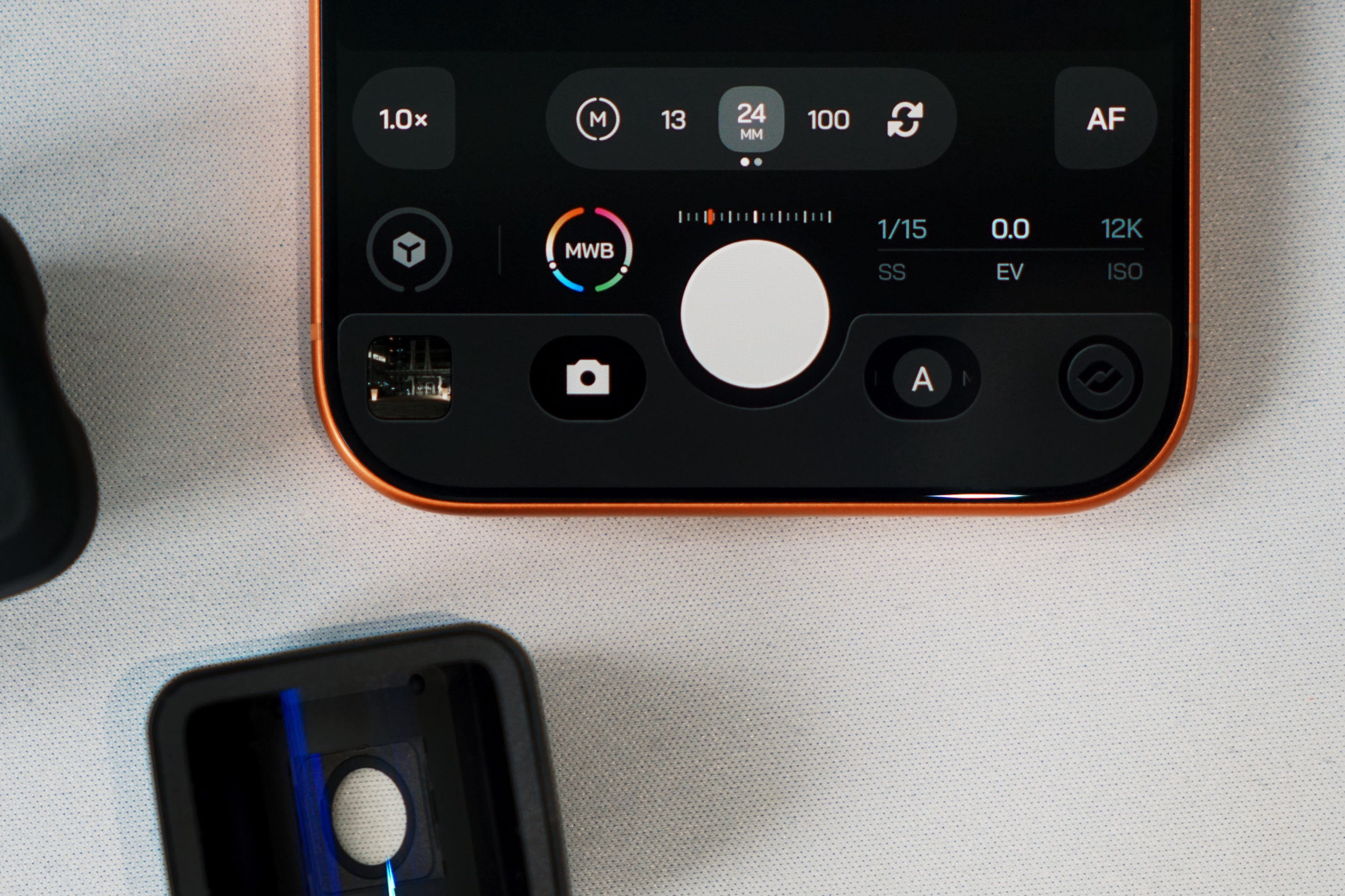 Close-up shot of a smartphone screen displaying a camera interface. The screen has a black background with several icons and text elements, including: "1.0x", "M", "13", "24 MM", "100", a refresh icon, "AF", "MWB", a shutter speed indicator showing "1/15 SS", an exposure value indicator showing "0.0 EV", and an ISO indicator showing "12K ISO". Other buttons are present, along with a large white circle in the center. The phone has an orange border and sits on a textured white surface, with a black device partially visible in the bottom left.
