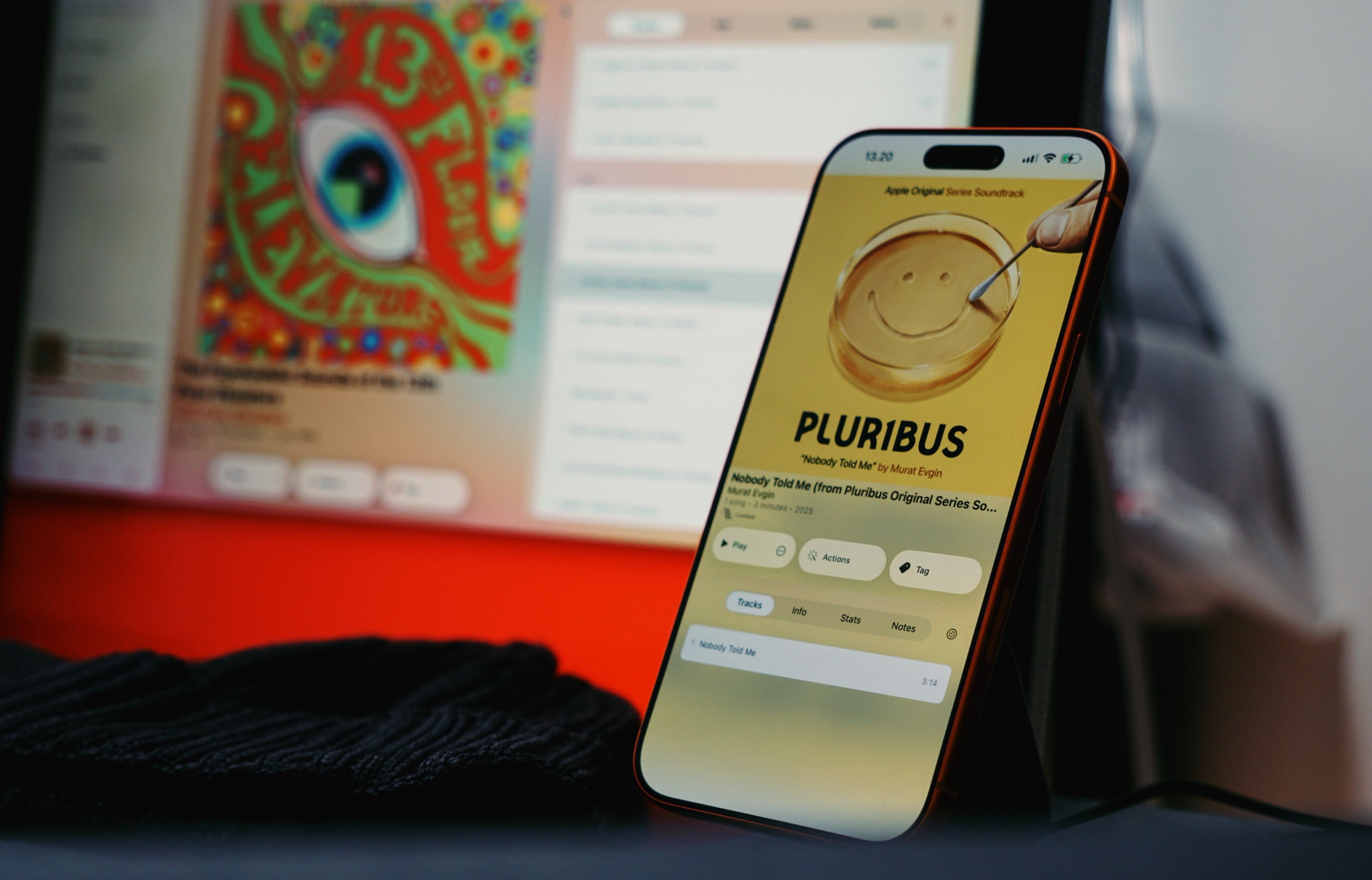 A close-up shot of a smartphone in front of a computer screen, with a blurred colorful image of "13 FLOOR" visible on the computer screen. The smartphone displays an Apple Music playlist page, featuring the song "Nobody Told Me" by Murat Evgin from the "Pluribus" soundtrack, with the text "PLURIBUS". The album art is a petri dish with a smiley face drawn in the center. The time, "13:20", is shown at the top of the smartphone screen.
