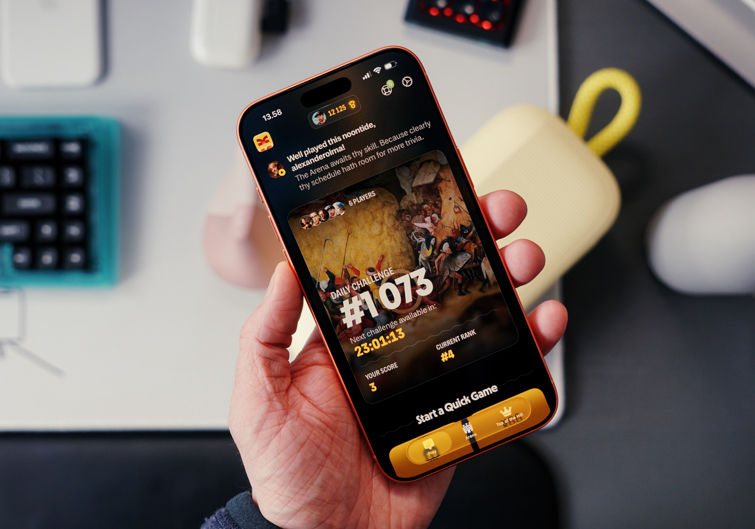 A hand holding an orange iPhone displaying a trivia game app; the screen shows a daily challenge with a game image, score, rank, and a countdown timer.

German Translation:
Eine Hand, die ein oranges iPhone hält, das eine Trivia-Spiel-App anzeigt; der Bildschirm zeigt eine tägliche Herausforderung mit einem Spielbild, einer Punktzahl, einem Rang und einem Countdown-Timer.
