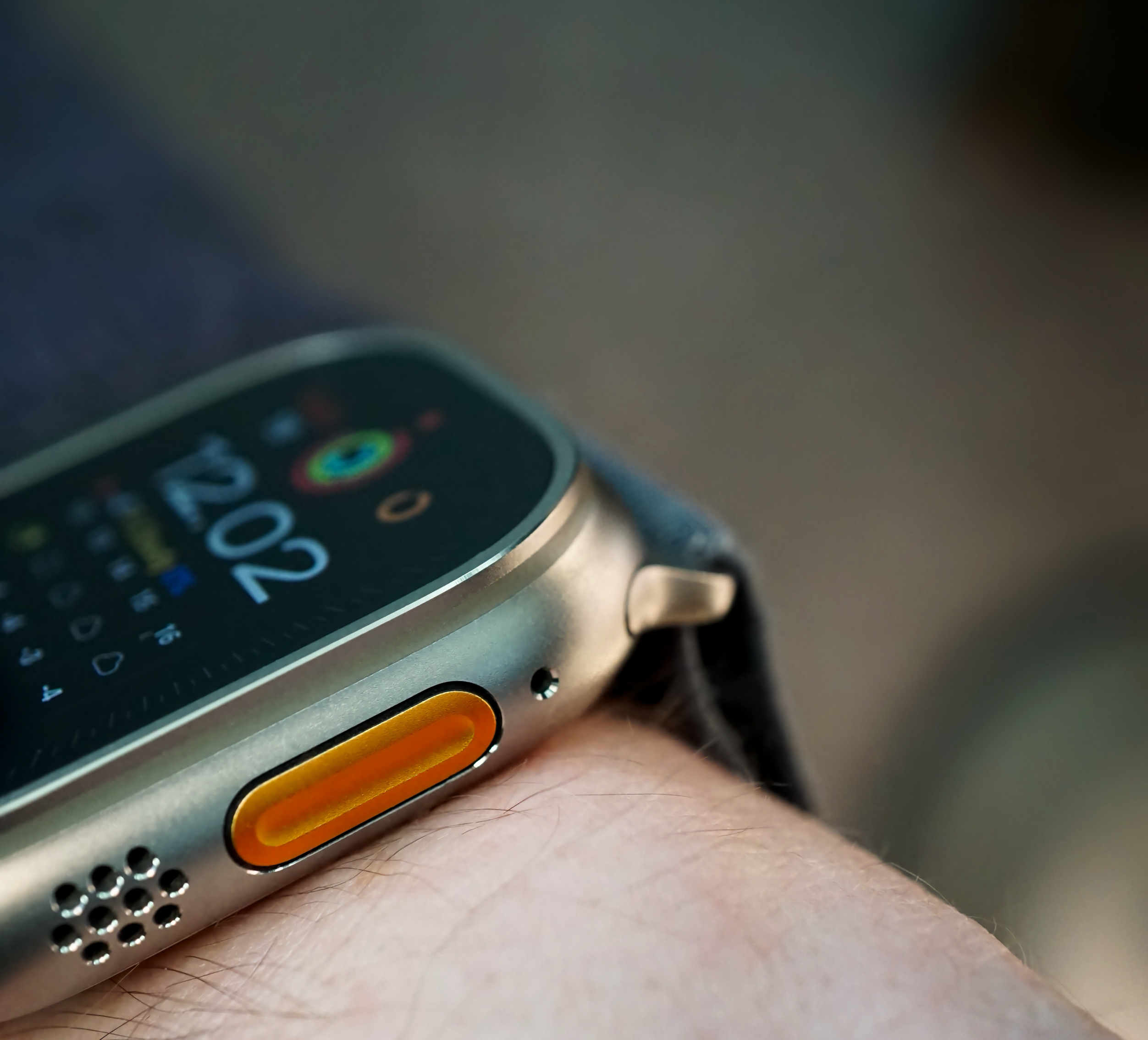 A close-up shot of an Apple Watch Ultra on a person's wrist. The watch has a silver titanium case with an orange button on the side. The watch face displays the time as "12:02". The strap is a dark gray color.