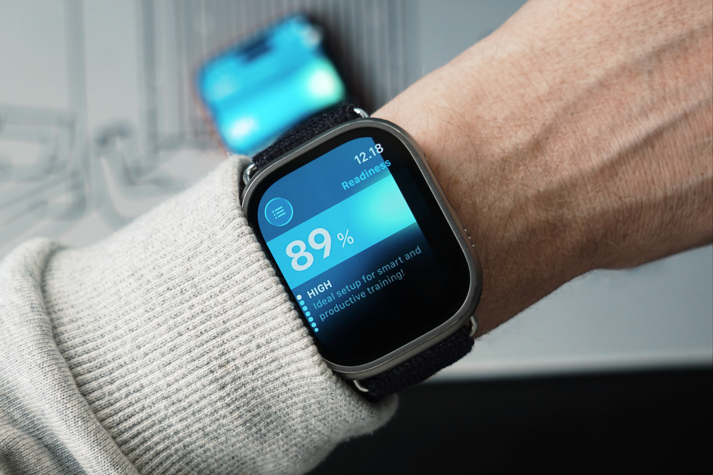 A wrist-worn smartwatch displaying fitness data. The screen displays the time "12.18" and the word "Readiness" above a percentage readout of "89%". Below that, the word "HIGH" is displayed with the text "Ideal setup for smart and productive training!". The watch is worn on a wrist covered by a light gray sweater. Another blurry object is behind the watch.