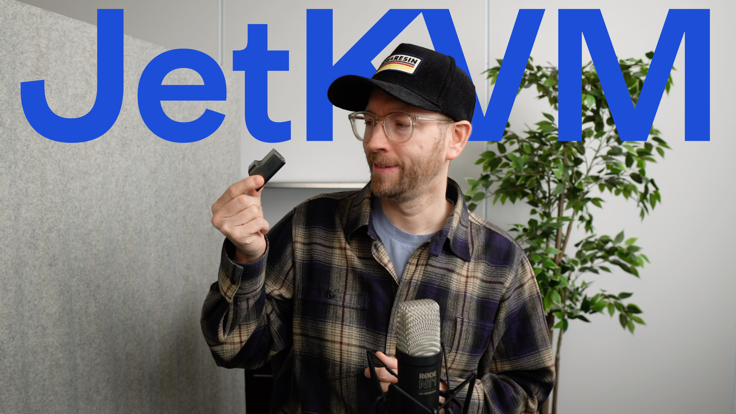 Ein Mann mit Brille und schwarzer Mütze, der ein kleines Gerät, wahrscheinlich ein Stück Technik, hält, mit "JetKVM" in großen blauen Buchstaben über ihm, ein Mikrofon vor sich.