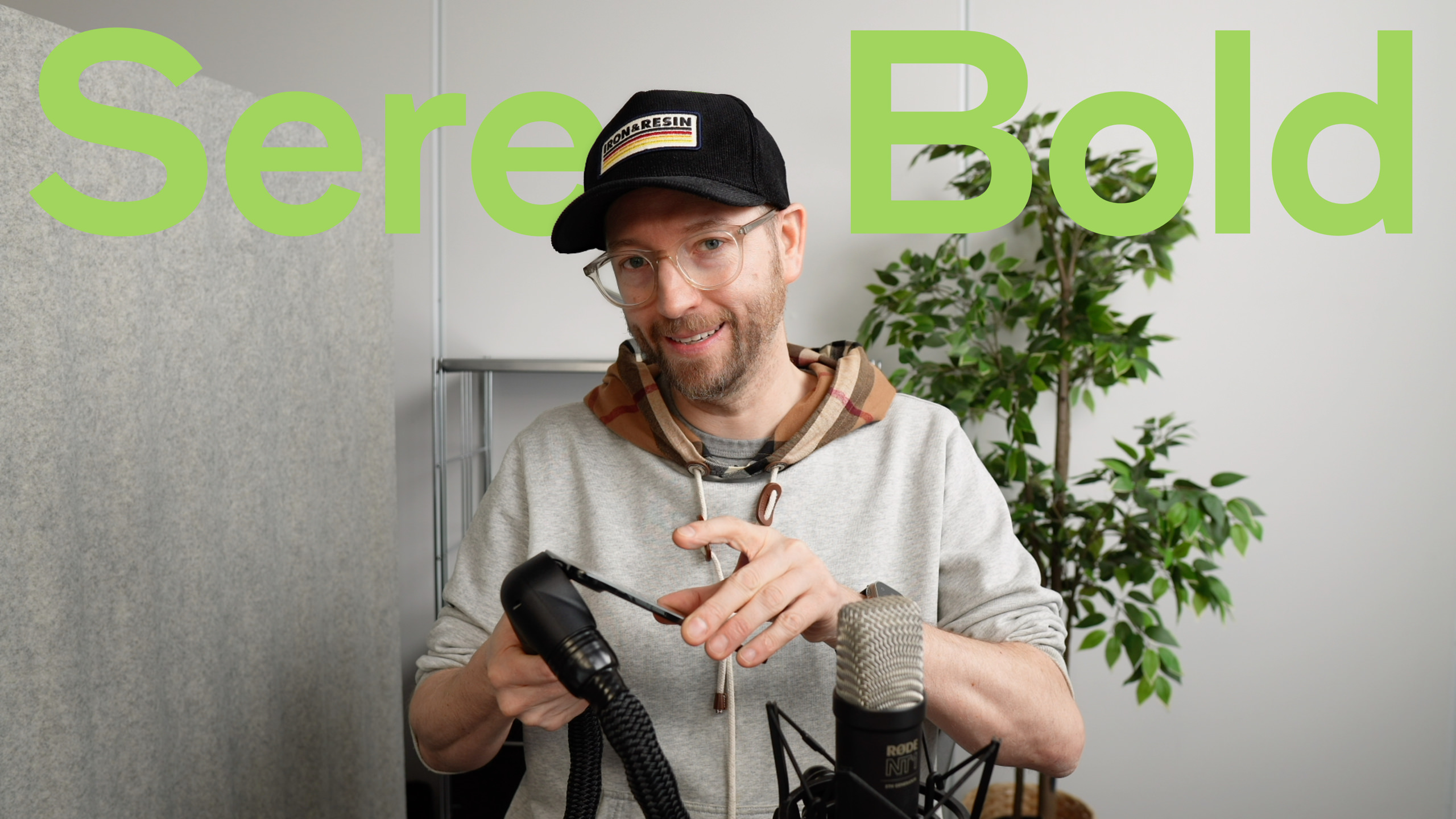 Mann mit Bart und Brille, der eine Mütze und einen Kapuzenpullover trägt, hält ein Mikrofonzubehör und lächelt in die Kamera in einem Studio.