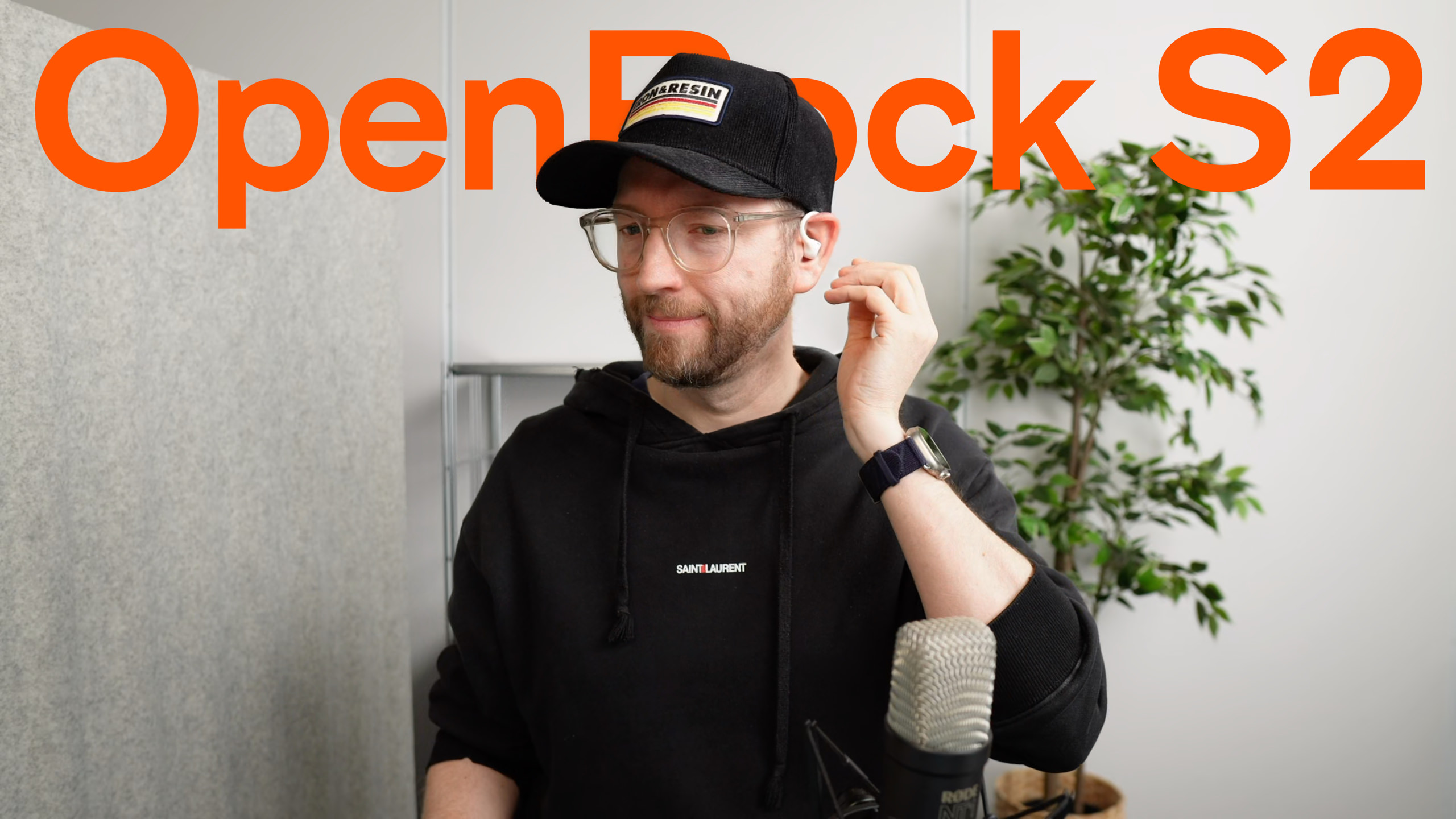 Ein Mann mit schwarzer Kappe, Brille und Kapuzenpullover, der einen Ohrhörer einstellt, mit den in orangefarbenen Buchstaben geschriebenen Worten "OpenRock S2" im Hintergrund.