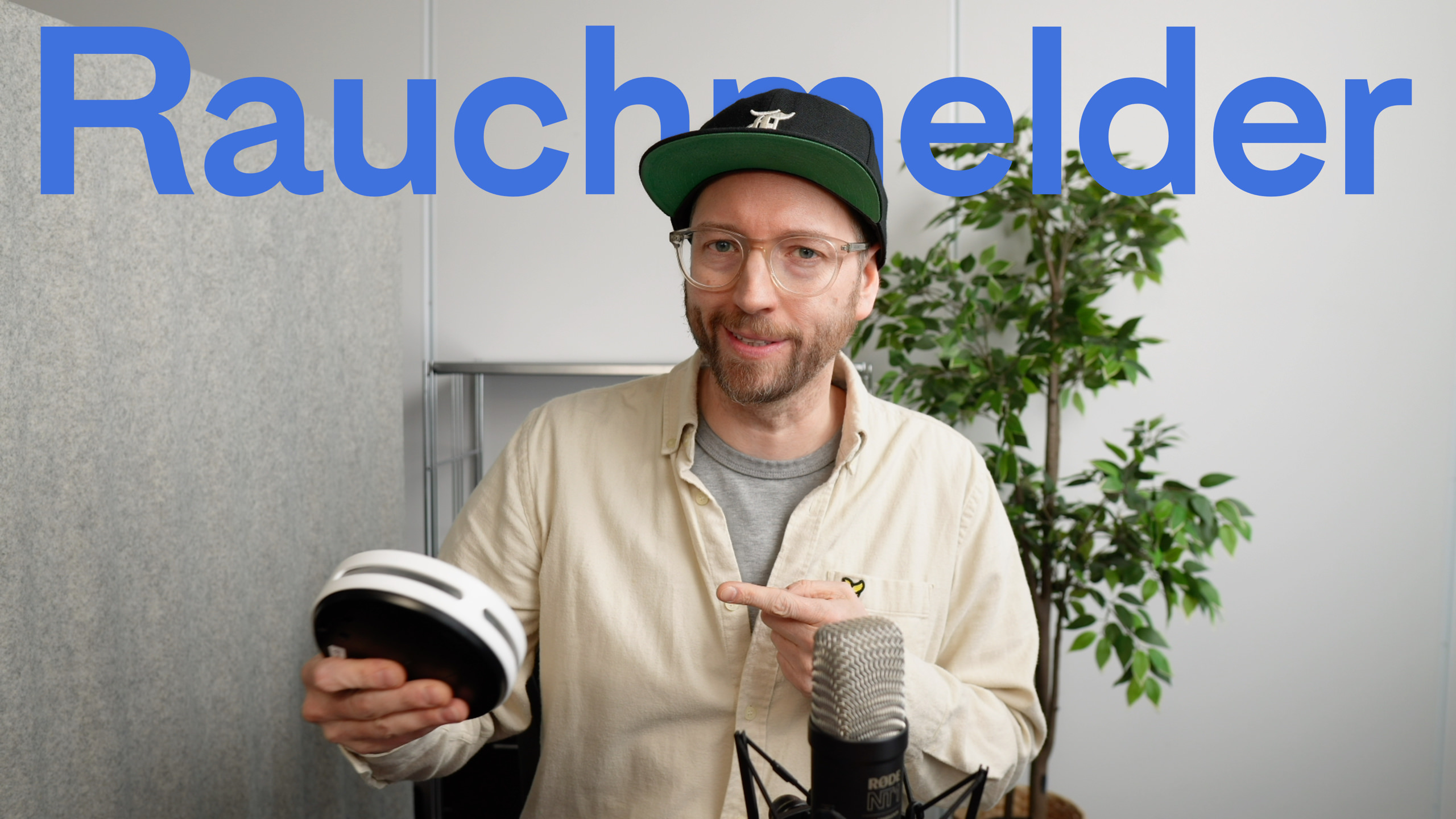 Mann mit Mütze und Brille hält einen Rauchmelder und zeigt auf das Gerät mit dem Text "Rauchmelder" in blauen Buchstaben über ihm.