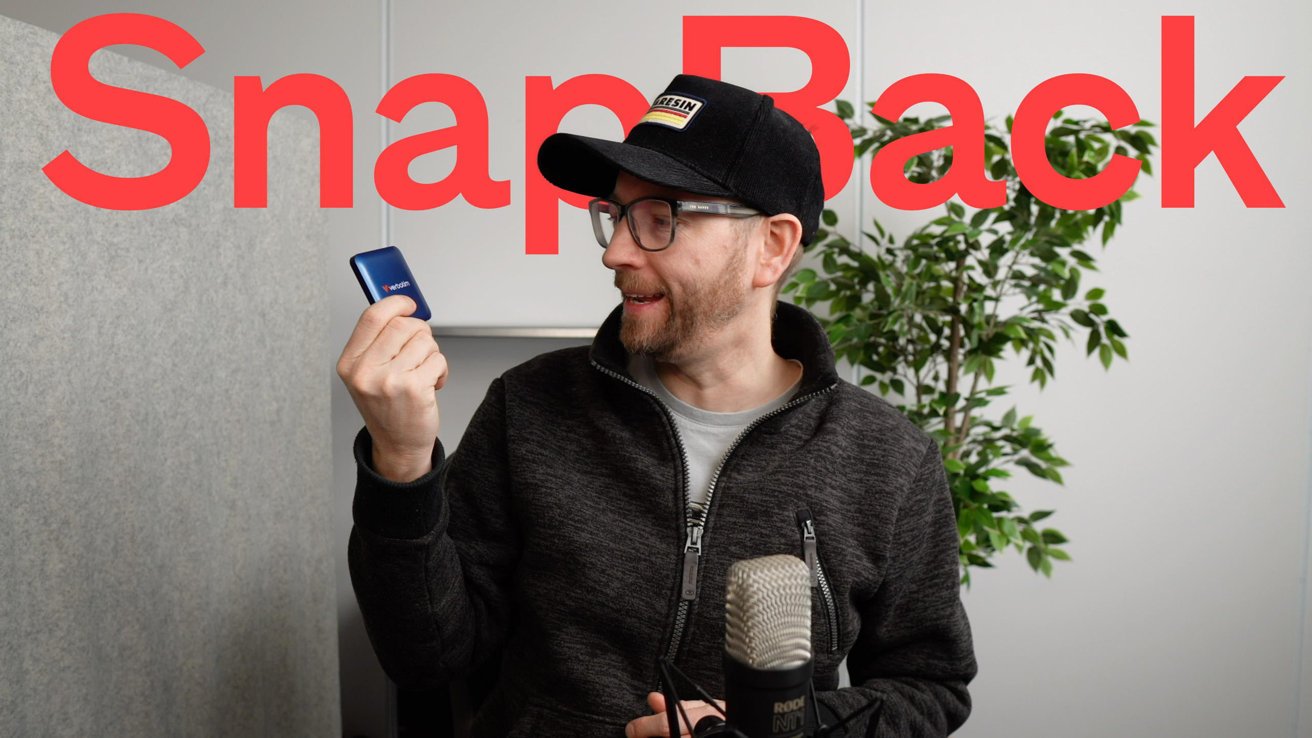 Ein Mann mit Brille und Hut hält eine blaue externe Verbatim-Festplatte vor einem Hintergrund, der einen roten "SnapBack"-Titel und ein Mikrofon enthält.

