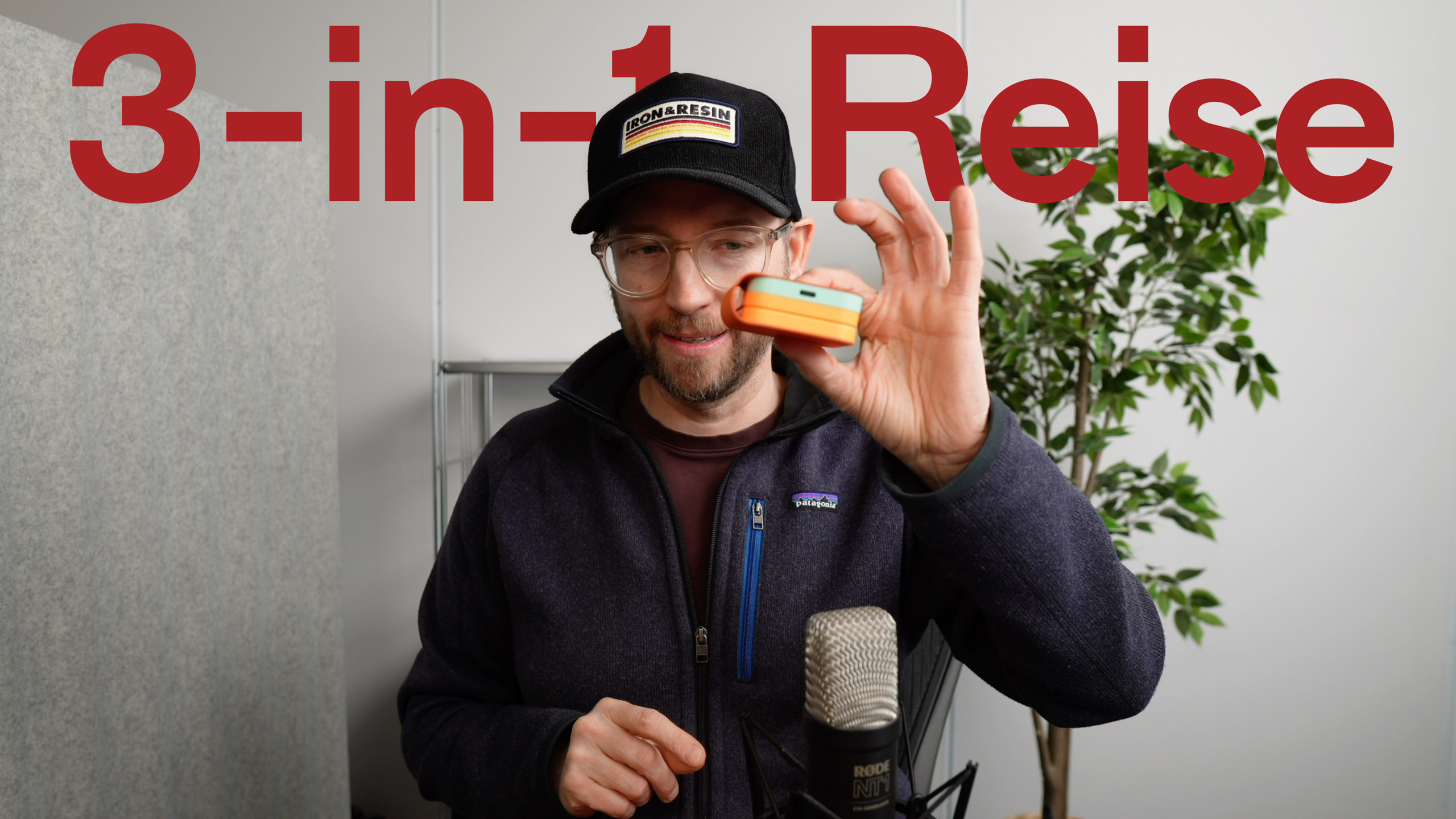 Mann mit Brille und Hut, der ein kleines orangefarbenes und teal-farbenes technisches Gerät hält, mit den Worten "3-in-1 Reise" in großen roten Buchstaben darüber, wahrscheinlich überprüft er das Produkt.