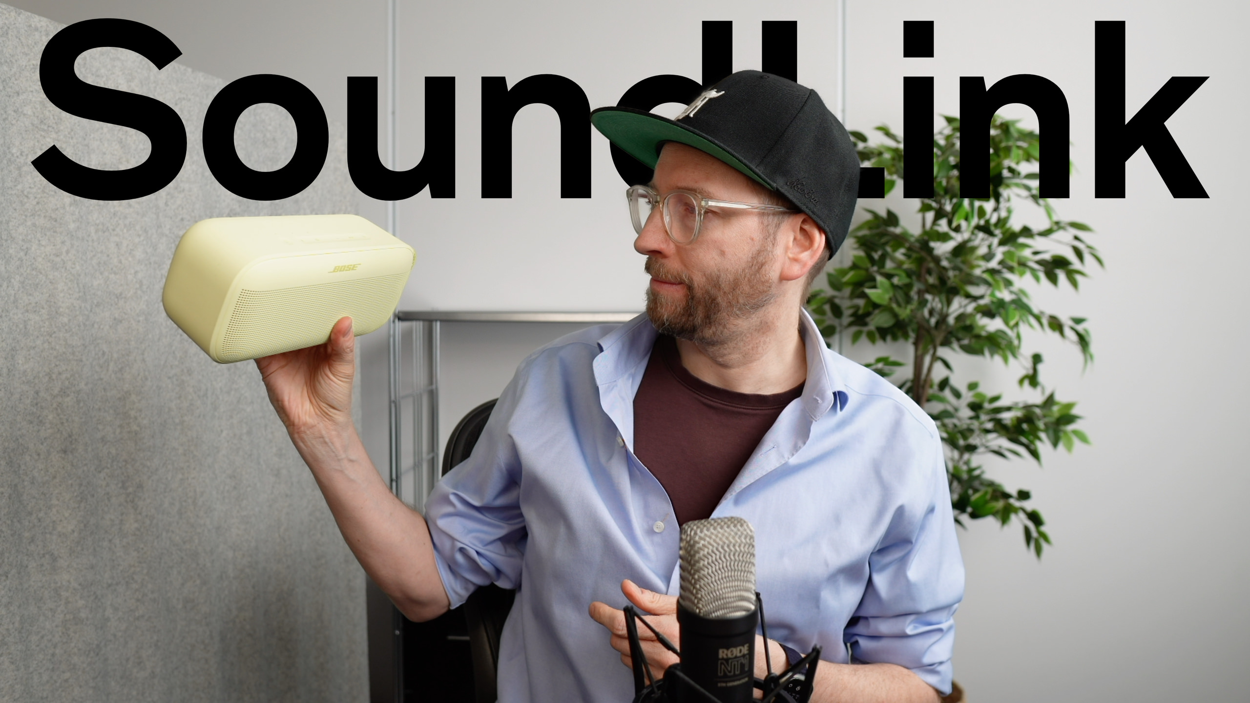 Mann mit Brille und Kappe, der einen gelben Bose Soundlink Flex-Lautsprecher hält und anscheinend eine Rezension oder Präsentation aufzeichnet, mit dem Wort "Soundlink" im Hintergrund.
