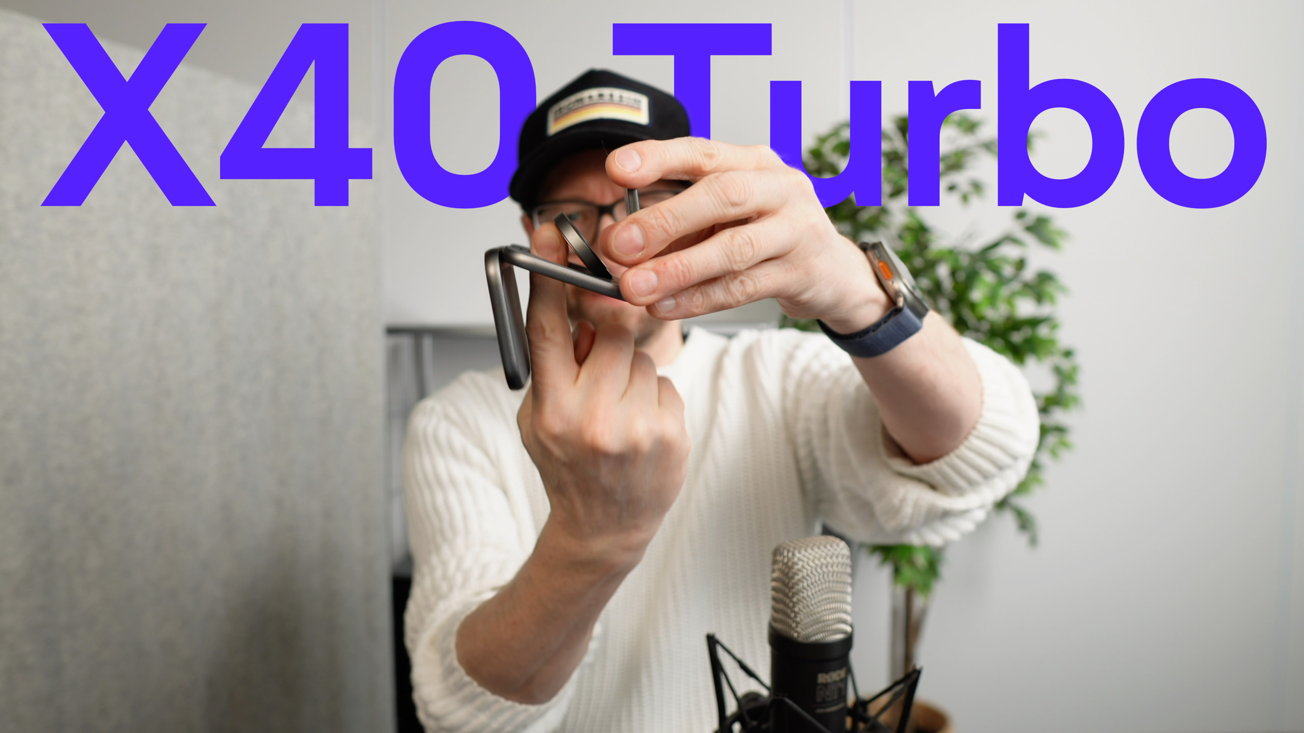 Eine Person mit schwarzer Mütze und Brille hält einen kleinen, schwarzen Metallgegenstand vor sich und demonstriert ihn scheinbar. Der Text "X40 Turbo" erscheint über der Person.