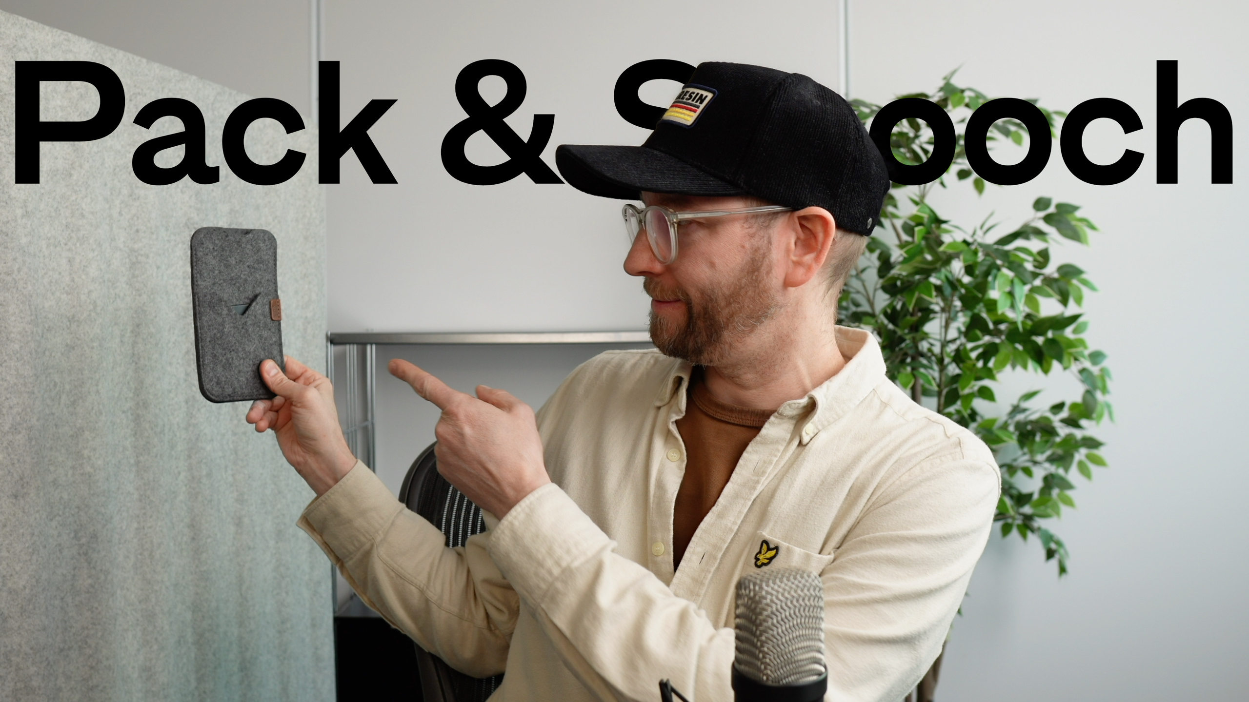 Ein Mann mit einer schwarzen Baseballkappe und Brille, der auf eine graue Filz-Handyhülle zeigt, die er hält, mit den Worten "Pack & Smooch" in großen schwarzen Buchstaben über ihm.