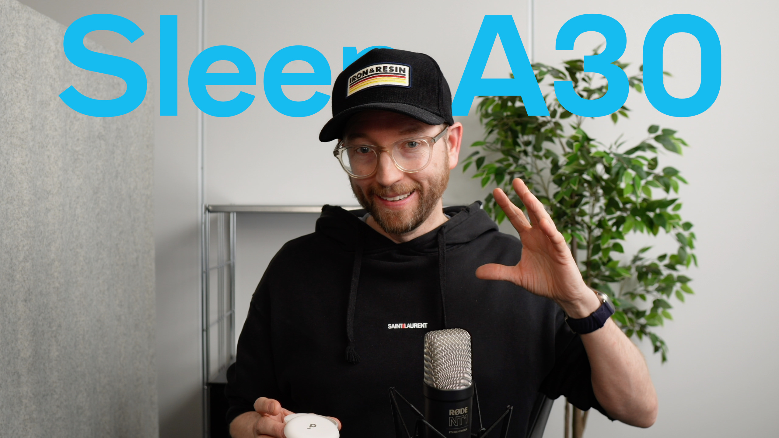 
Mann in schwarzem Kapuzenpullover und Mütze mit "Iron & Resin"-Aufnäher und klarer Brille, hält einen weißen, abgerundeten Gegenstand und gestikuliert mit der anderen Hand, mit dem Wort "Sleep A30" in großen tealblauen Buchstaben über ihm; vor einem Mikrofon in einem Aufnahmestudio.
