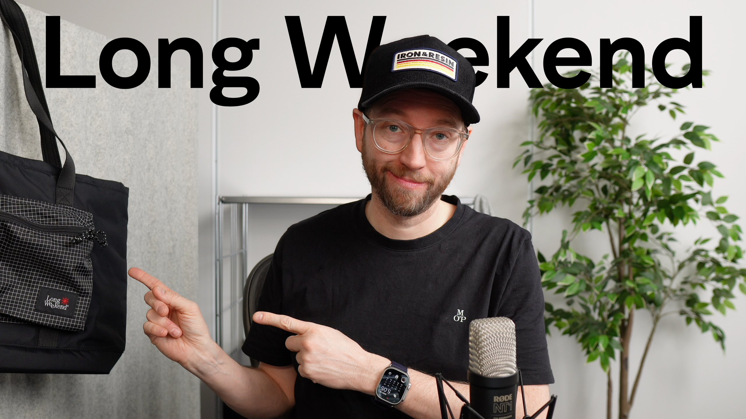 Mann mit Brille und Kappe, der auf eine Tragetasche und ein Mikrofon zeigt, mit den Worten "Long Weekend" über ihm.
