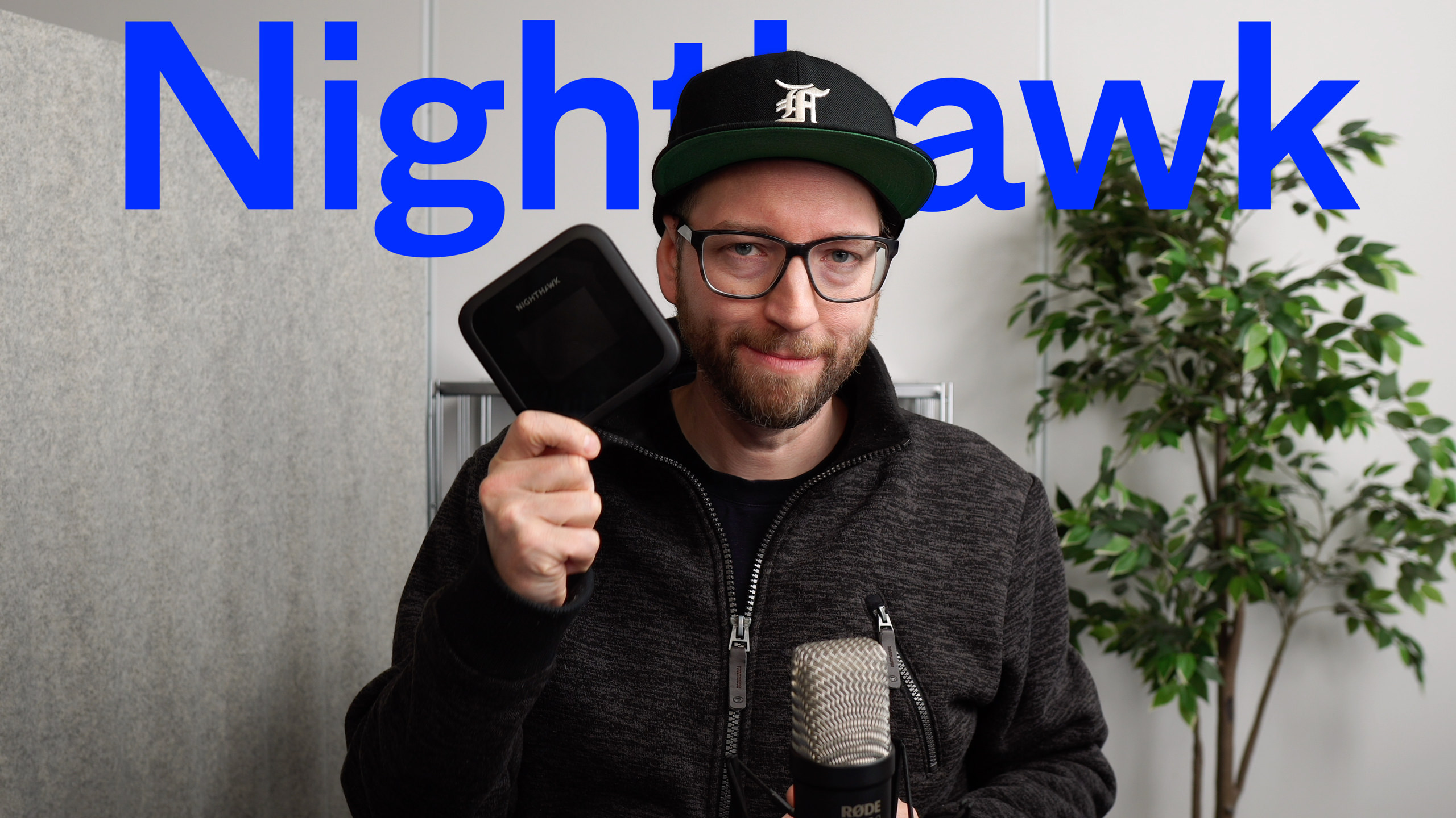 Mann mit Brille und Hut hält ein schwarzes Nighthawk-Gerät unter einem blauen "Nighthawk"-Text hoch, in der Nähe eines Mikrofons und einer künstlichen Pflanze.
