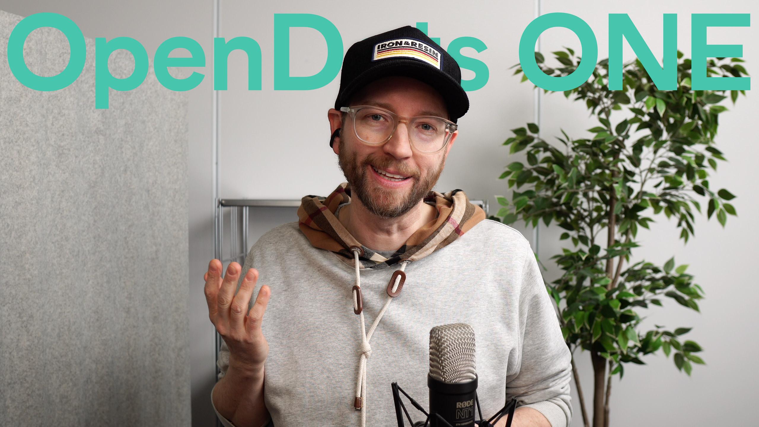 Ein lächelnder Mann mit Brille, Hut und Kapuzenpulli steht vor einem Mikrofon, mit "OpenDots ONE" in Türkis über ihm geschrieben.