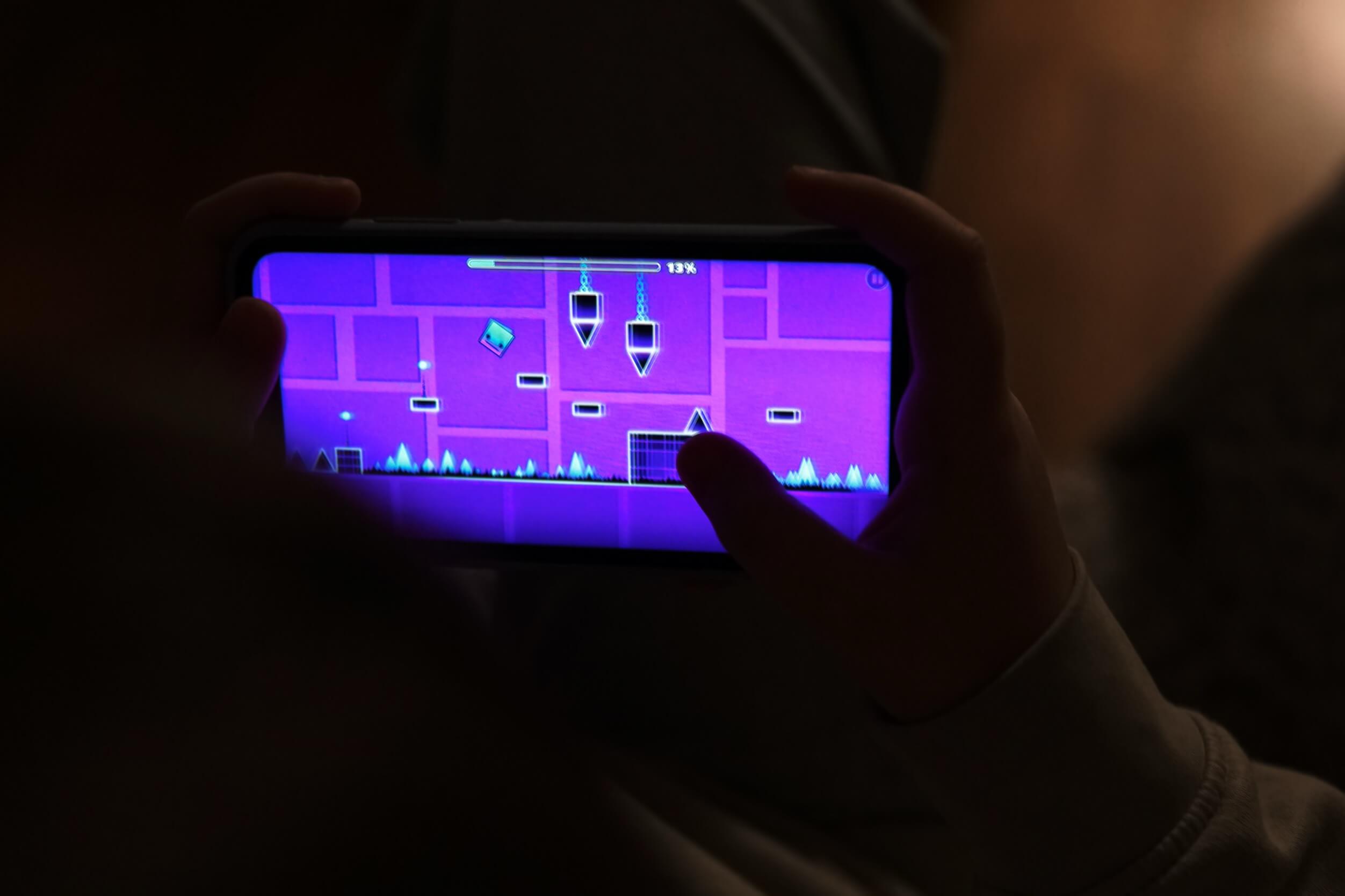 Eine Person hält ein Telefon und spielt ein Videospiel. Der Spielbildschirm zeigt ein geometrisches Level in Lila und Pink mit einem blockartigen Charakter, Blöcken und Stacheln. Das Spiel zeigt den Text "13%".
