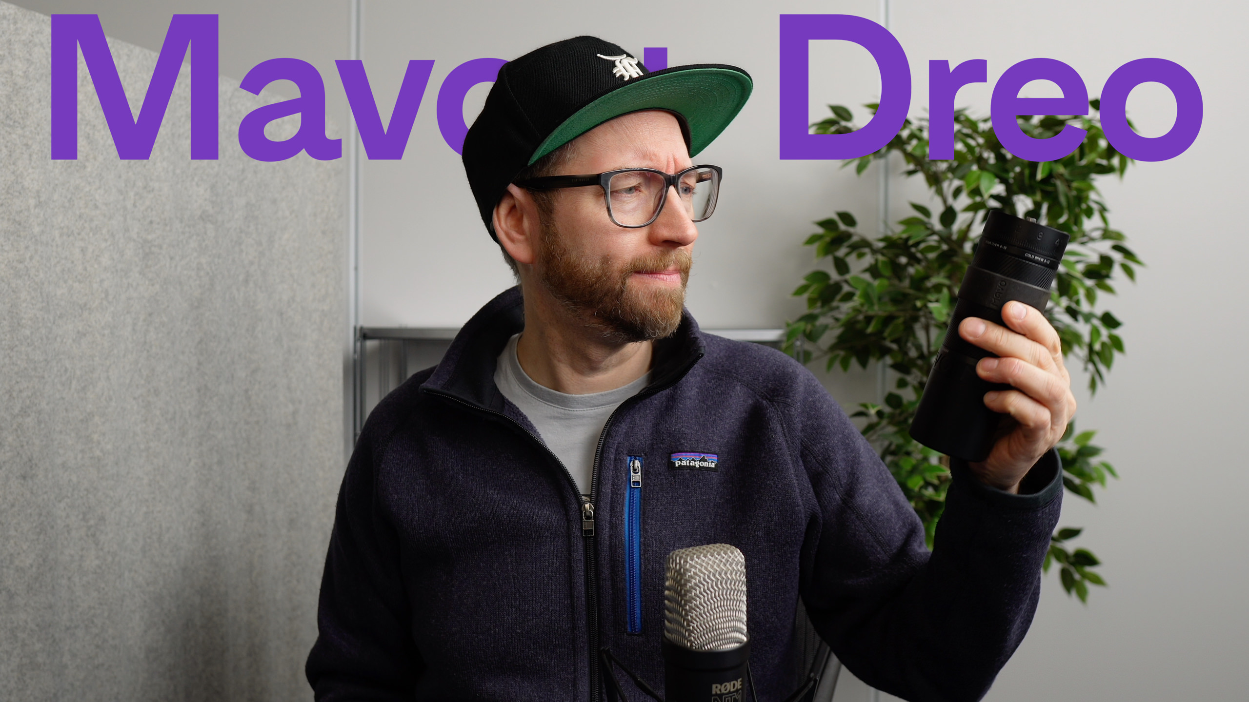 Ein Mann mit schwarzer Kappe, Brille und blauer Patagonia-Jacke betrachtet ein schwarzes zylindrisches Objekt, das er in seiner rechten Hand hält, vor einem neutralfarbenen Hintergrund mit den Worten "Mavo + Dreo" in Lila.
