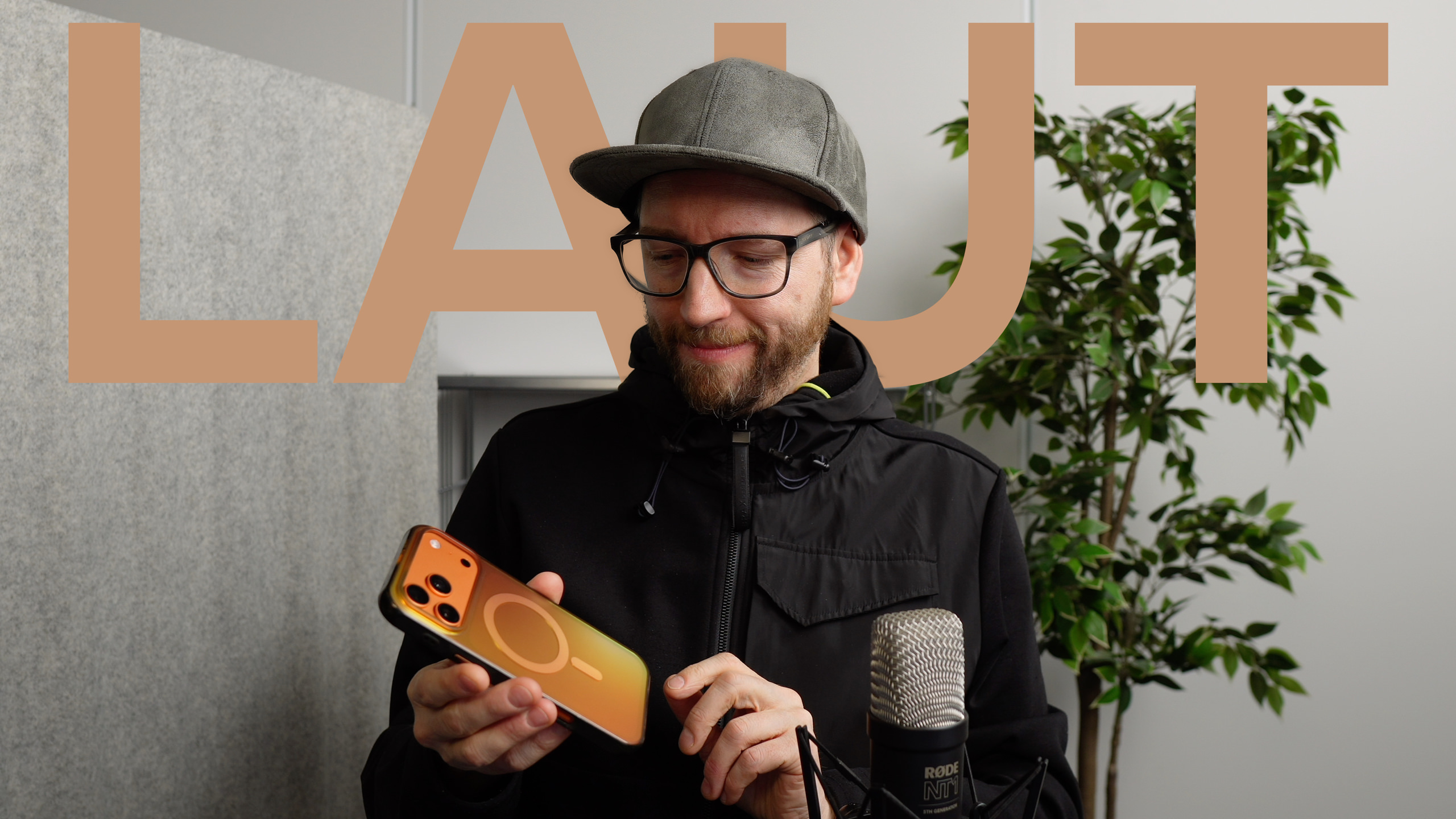 Ein Mann mit Brille und Mütze, der ein iPhone mit einer Hülle in Orangetönen hält und betrachtet. Das Wort "LAUT" in großen, durchscheinenden Buchstaben ist überlagert, mit einem Mikrofon und einer Topfpflanze im Hintergrund.
