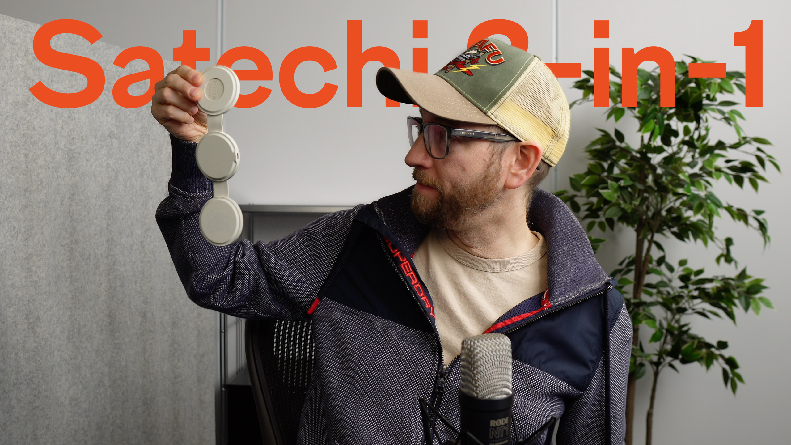 Ein Mann mit Brille und Trucker-Hut hält ein weißes, dreiteiliges Audiogerät vor dem Hintergrund des orangefarbenen Textes "Satechi 3-in-1" hoch.