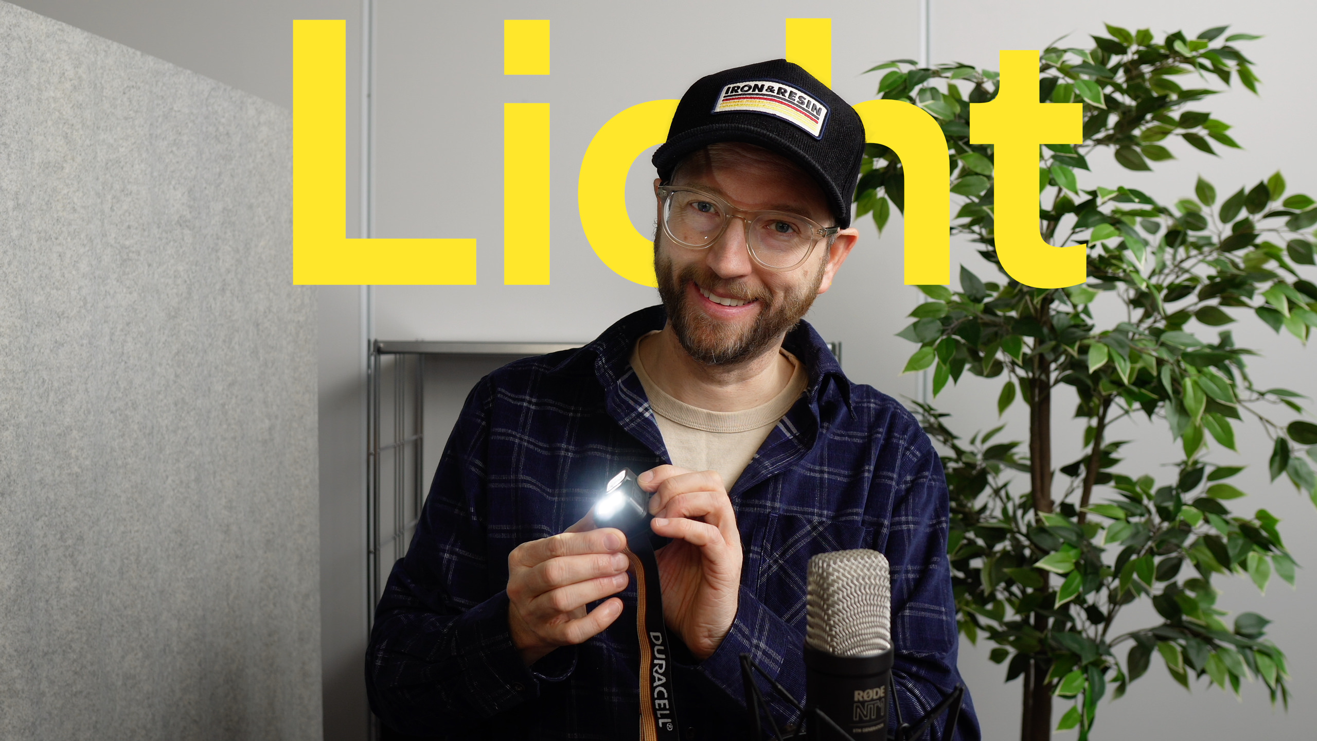 Ein lächelnder Mann mit schwarzer Kappe und Brille hält eine Taschenlampe und hebt ihr Licht mit dem Wort "Light" in großen gelben Buchstaben im Hintergrund hervor.
