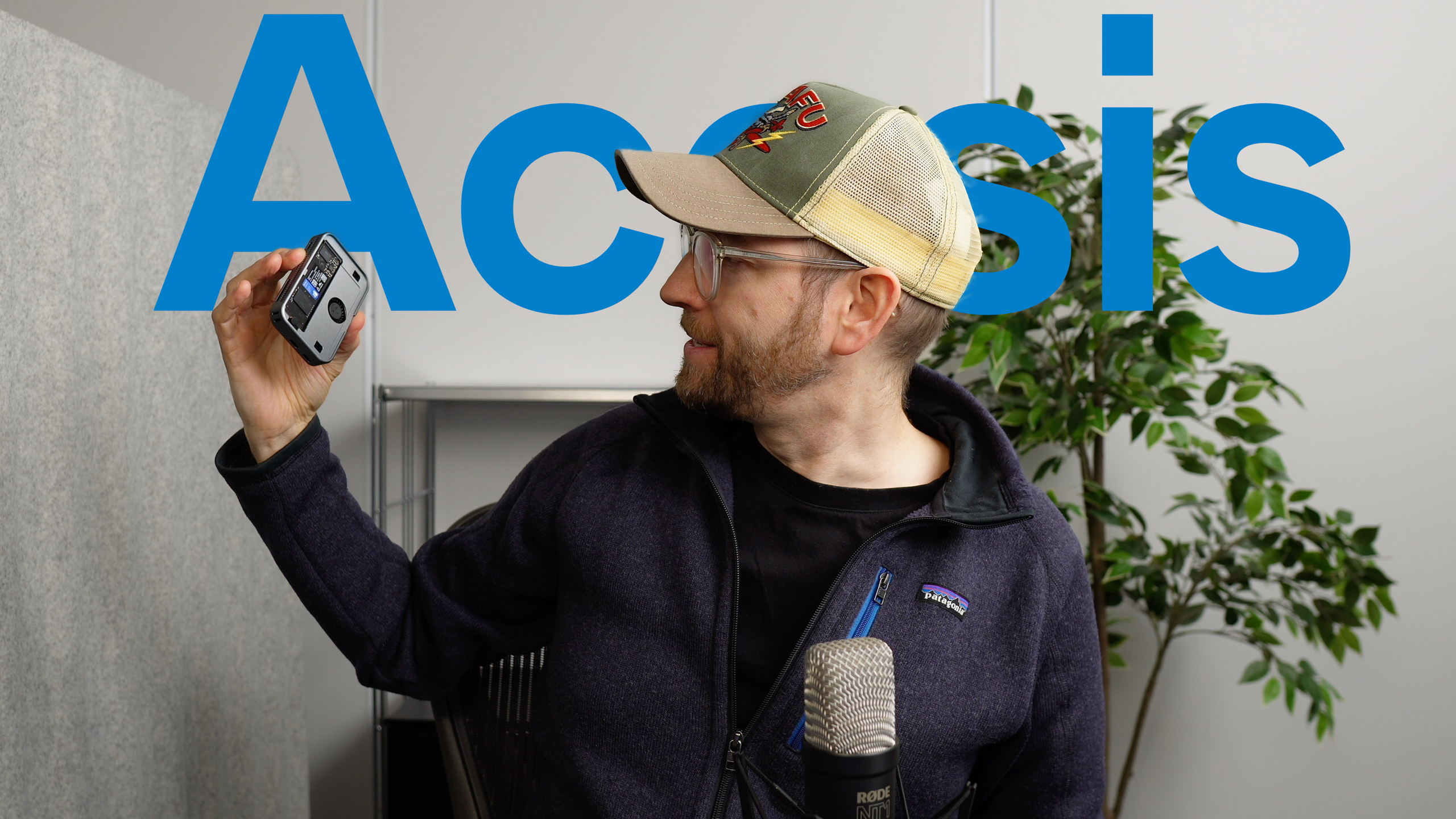 Ein Mann mit Brille und Trucker-Hut hält ein kleines silbernes Gerät, wahrscheinlich eine Kamera oder ein Aufnahmegerät, vor einem blauen "Acasis"-Text-Hintergrund und ist in der Nähe eines Mikrofons positioniert, was auf ein Video- oder Podcast-Setup hindeutet.
