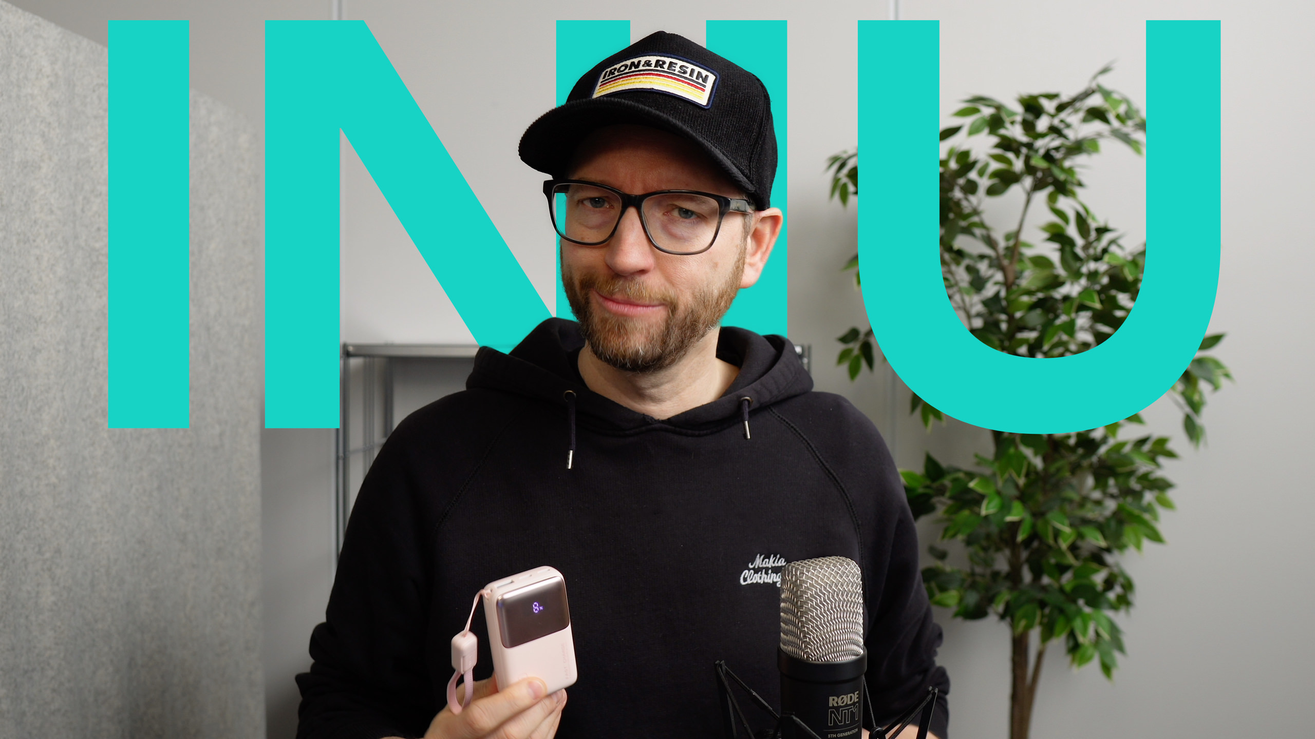 Ein Mann mit Bart und Brille, der eine Kappe und einen schwarzen Kapuzenpullover trägt und ein rosa Gerät hält, steht vor einer türkisfarbenen "INIU"-Grafik, wobei ein Mikrofon und eine Pflanze im Hintergrund sichtbar sind.