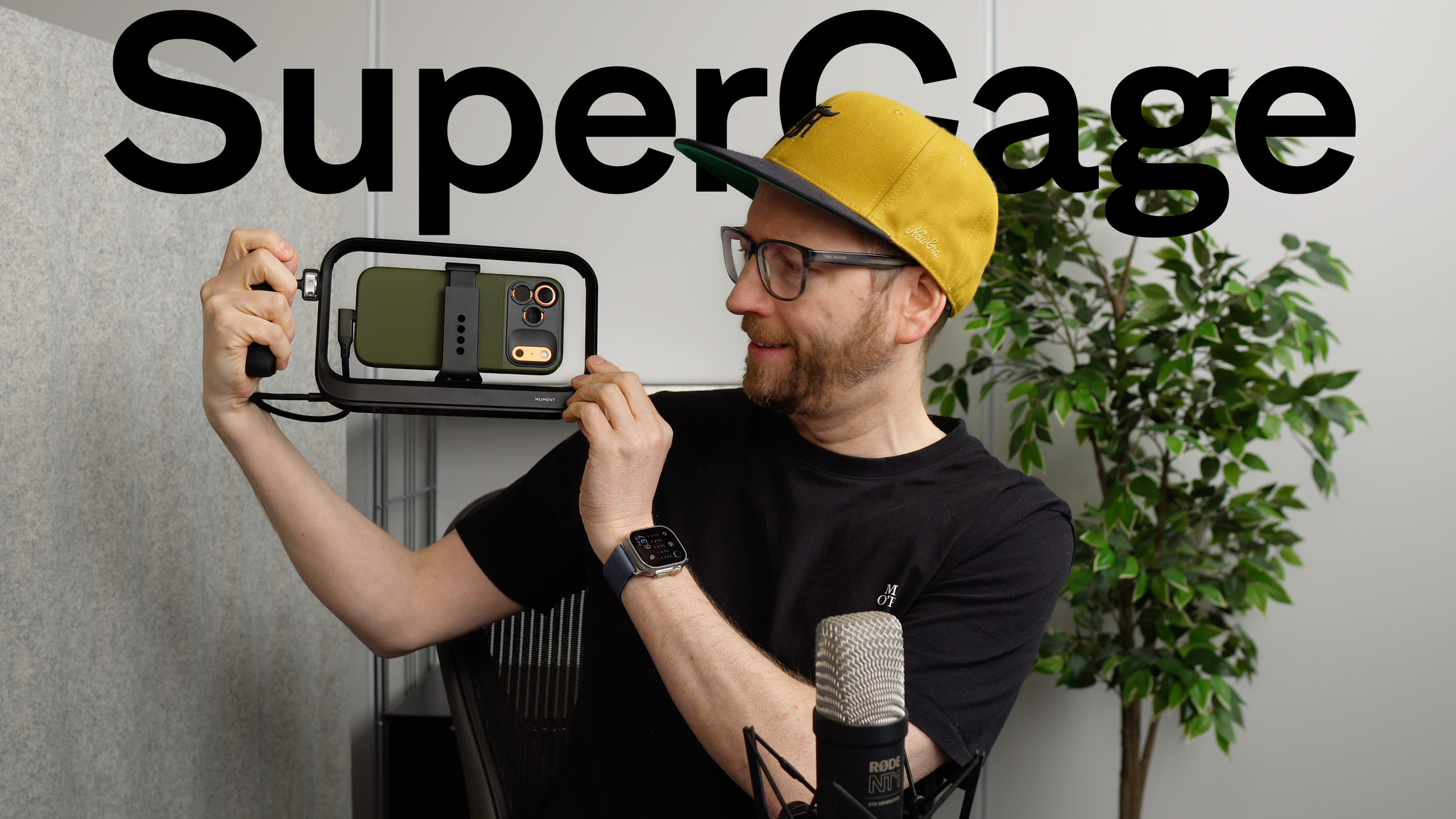 Mann mit Brille und gelber Mütze, lächelnd, hält eine schwarze Telefon-Cage mit einem olivgrünen Telefon darin, vor dem "SuperCage"-Logo, ein Mikrofon und eine Pflanze sind im Hintergrund.
