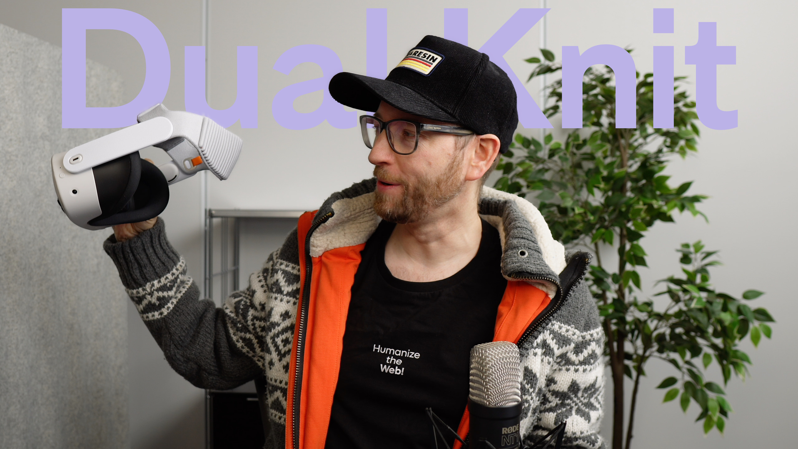 Ein Mann mit Brille und schwarzer Kappe hält ein weißes VR-Headset vor einem Hintergrund mit den Worten "Dual Knit", mit einem Mikrofon und einer Pflanze im Hintergrund.