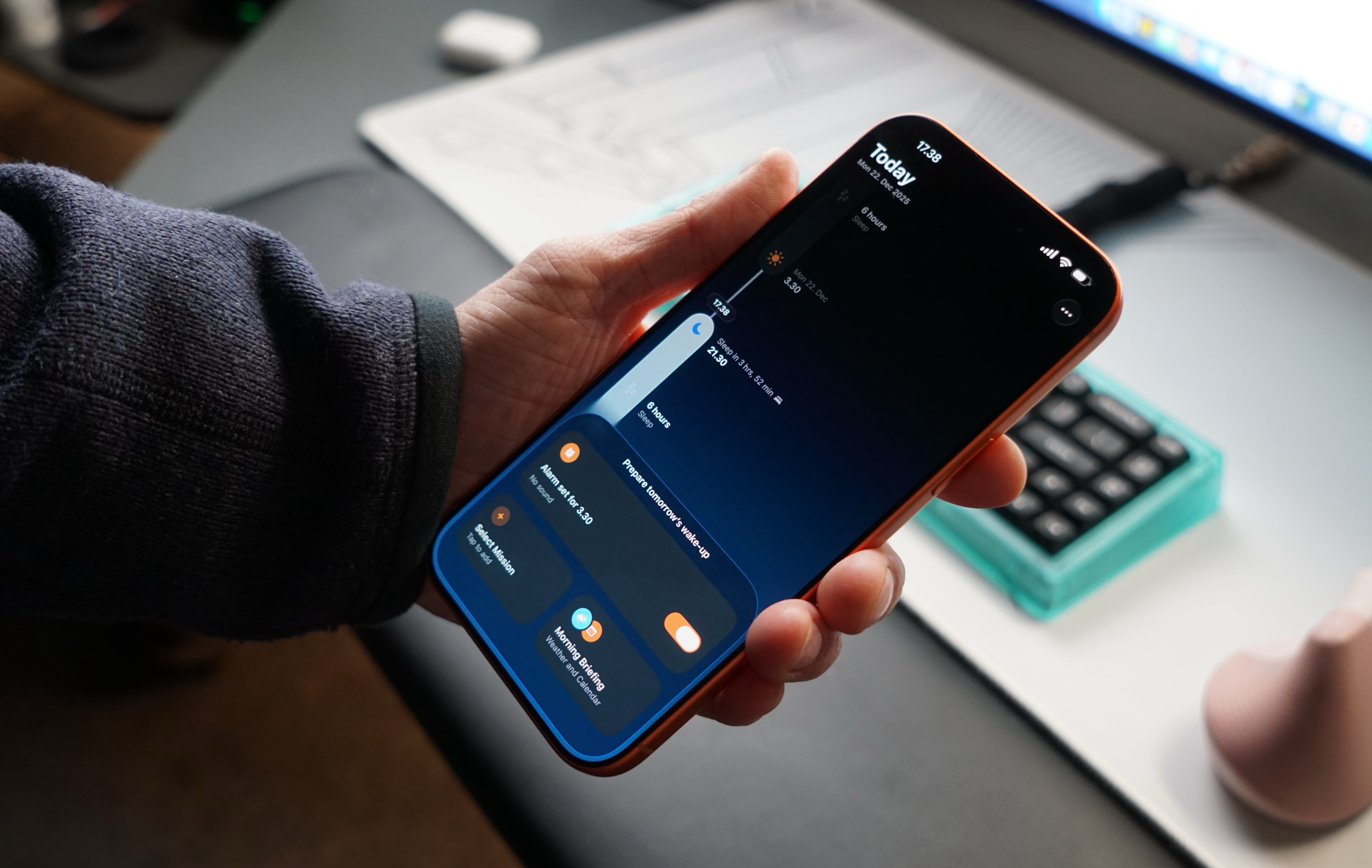 A hand holding an orange iPhone displaying the "Today" view in the clock app, showing sleep data, alarms, and the time 17:38.

German Translation:
Eine Hand, die ein orangefarbenes iPhone hält, auf dem die "Heute"-Ansicht in der Uhr-App angezeigt wird, mit Schlafinformationen, Weckern und der Uhrzeit 17:38 Uhr.
