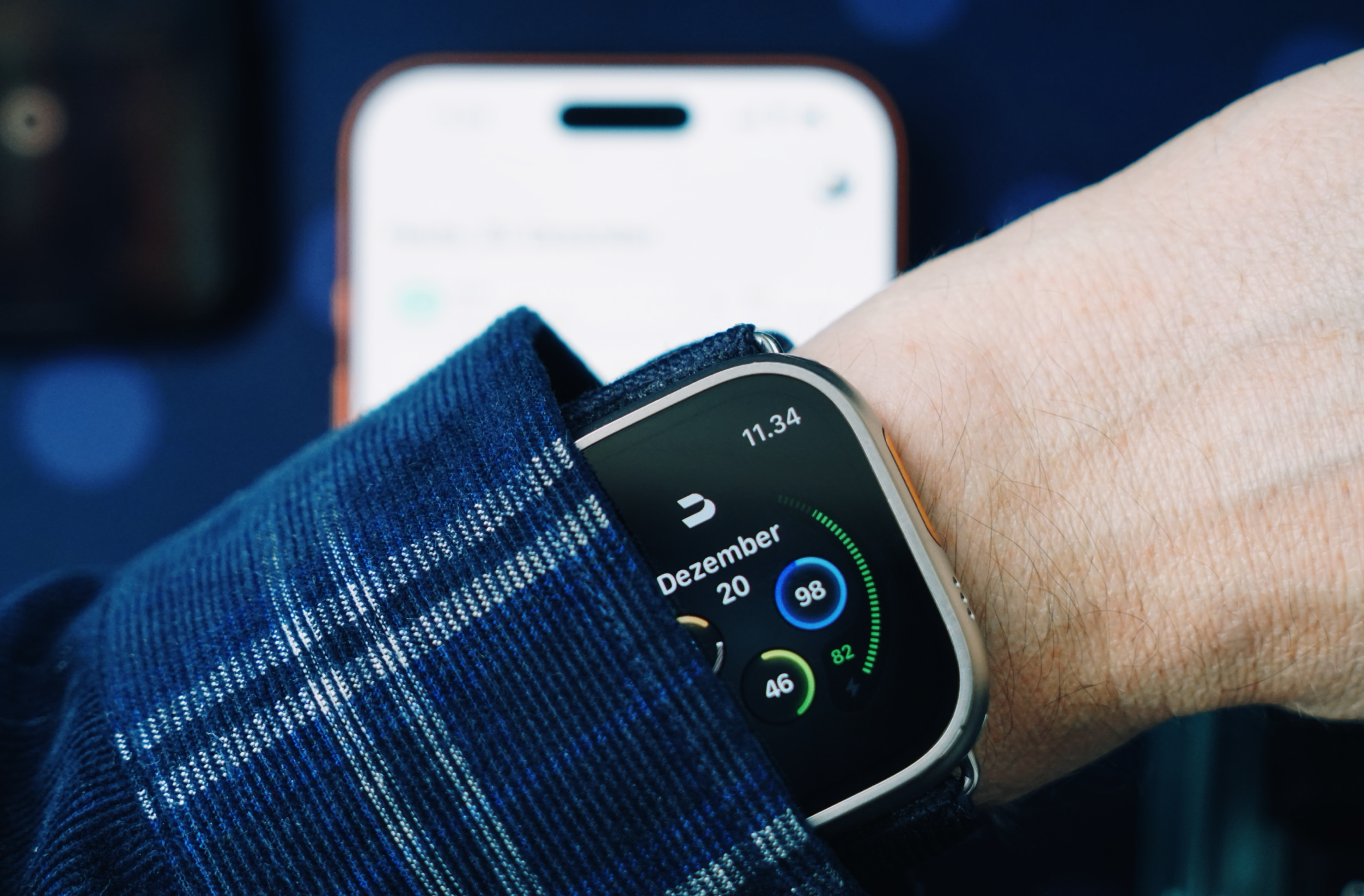 Eine Apple Watch Ultra, die Gesundheitsmetriken anzeigt, die an einem Handgelenk getragen wird, teilweise verdeckt durch ein blaues kariertes Hemd; ein verschwommenes Telefon ist im Hintergrund.
